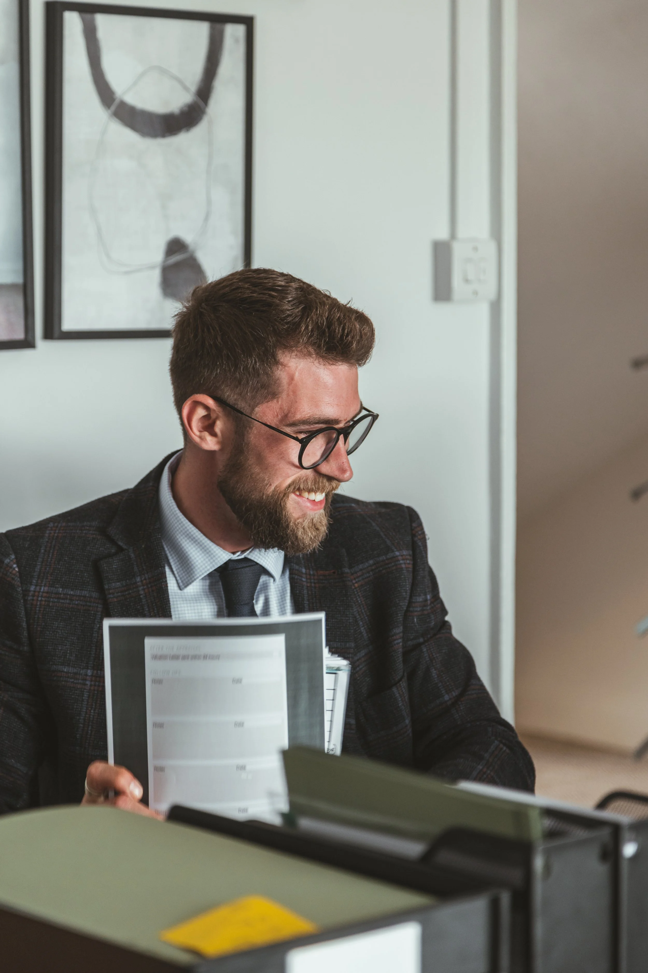 A smiling man with glasses, a beard, and short hair wearing a suit jacket, shirt, and tie, sitting at a desk in an office environment, holding some documents.