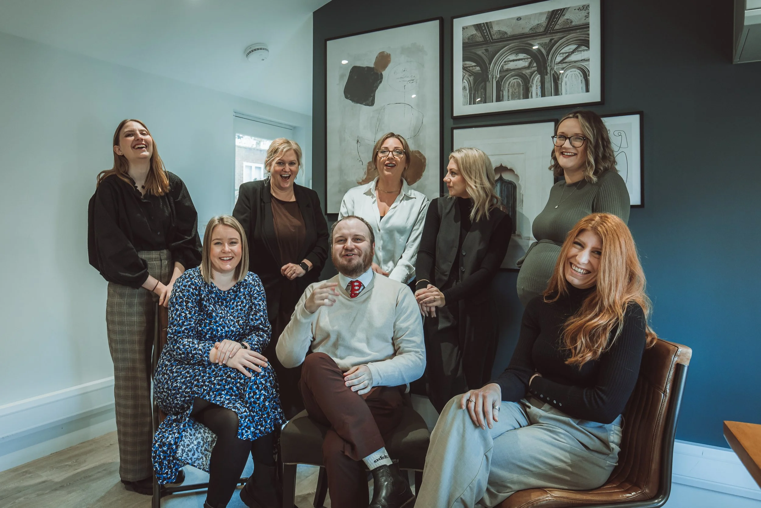 Group of nine people, consisting of seven women and two men, in a modern office or gallery space, smiling and laughing together. Behind them are framed abstract art and architectural photos on a gray wall.