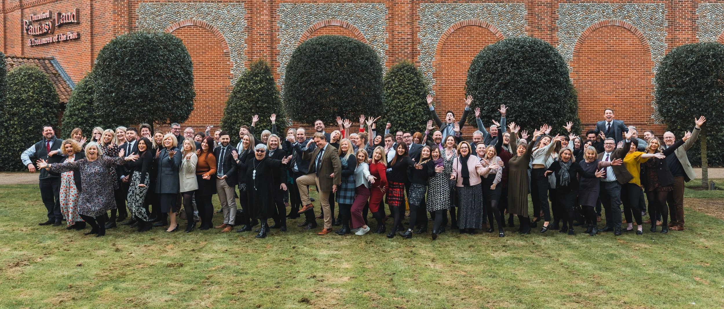 A large group of people in business attire posing and celebrating outdoors in front of a brick and stone building with trees.