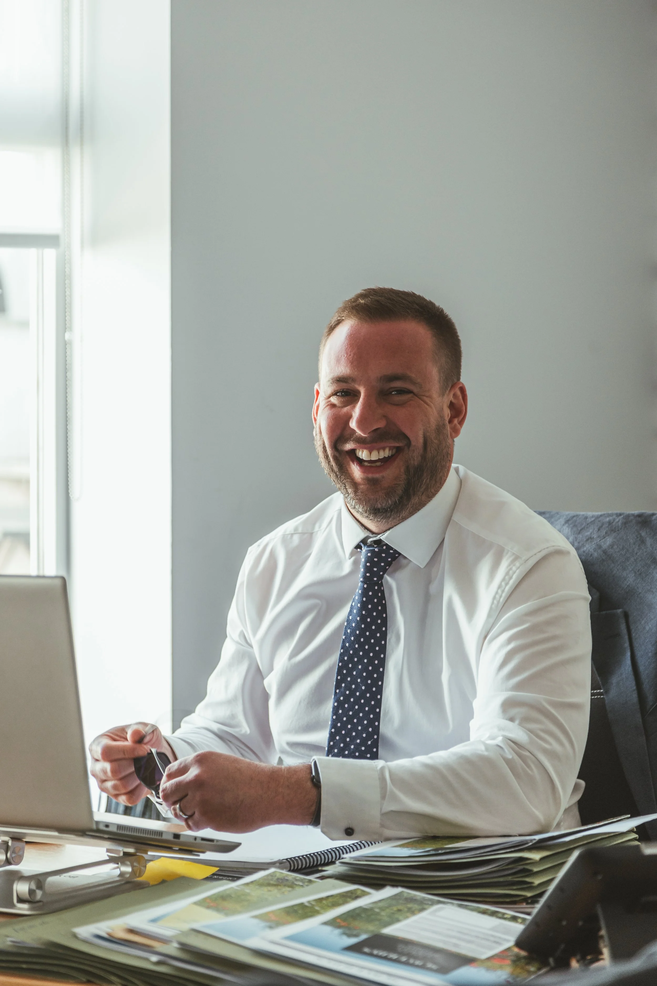 A man smiling and sitting at a desk in an office, with a laptop, papers, and magazines in front of him.