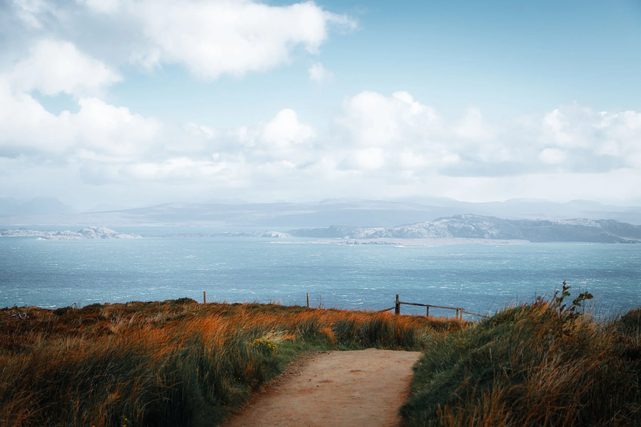 A dirt path leading toward the ocean with grassy dunes on either side, hills, and mountains in the distance under a partly cloudy sky.