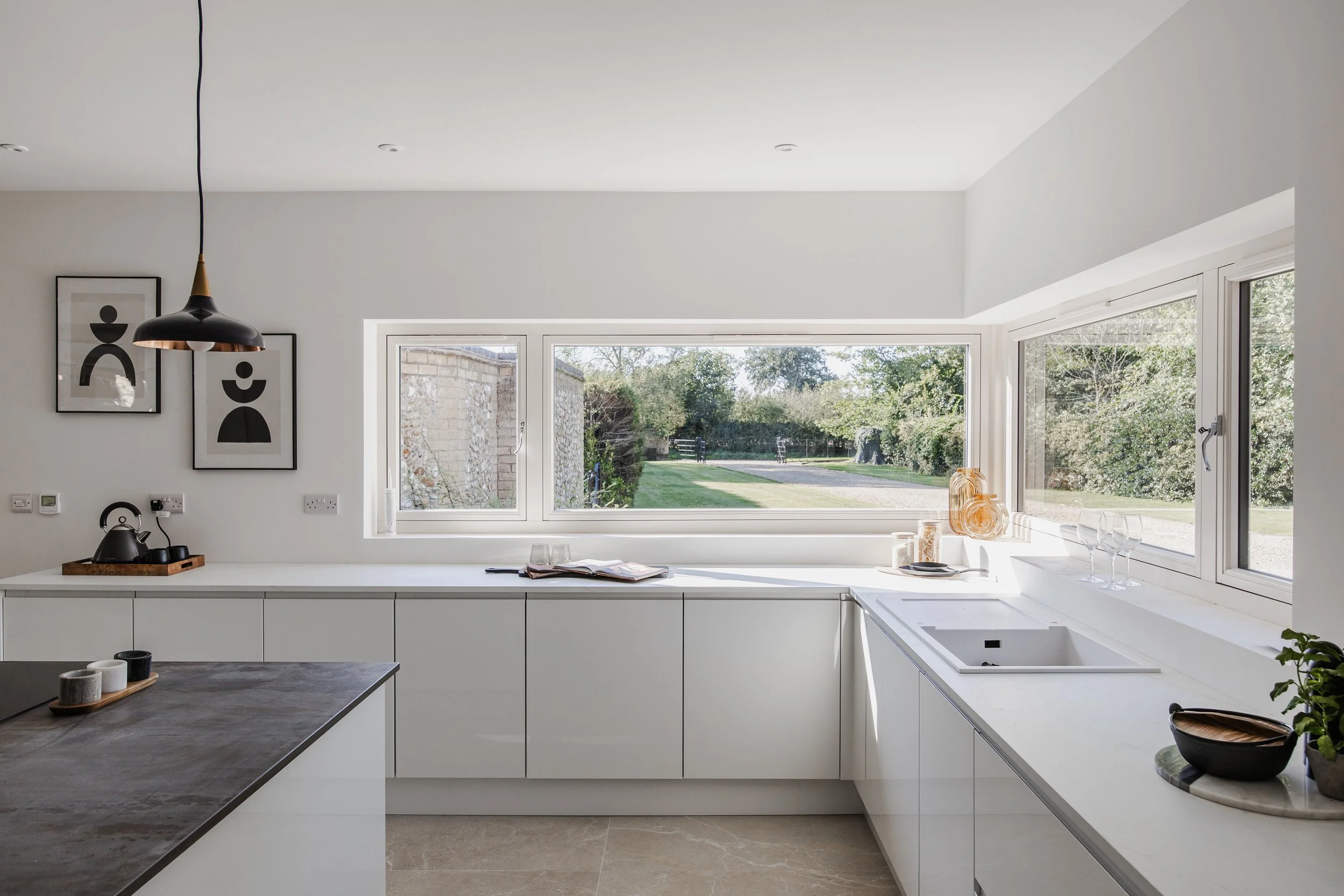 Modern kitchen with white cabinets, large windows showing a green outdoor scene, black and white artwork on the wall, and various kitchen items on the counters.