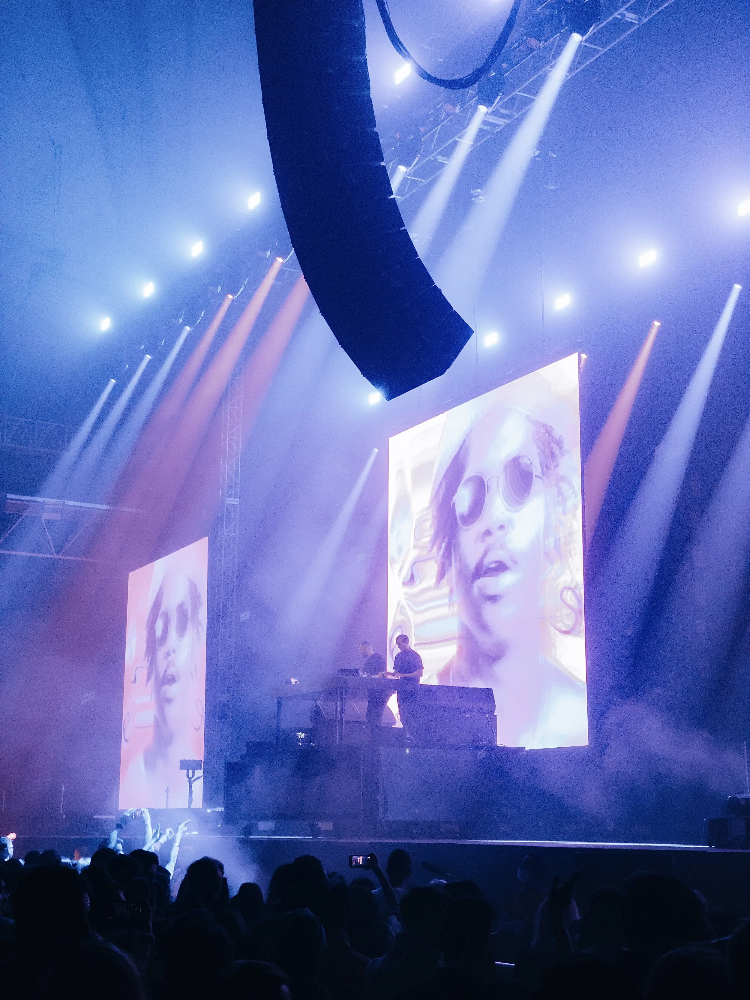A concert stage with LED screens displaying a person wearing sunglasses, surrounded by colorful light beams and a crowd of fans taking photos.
