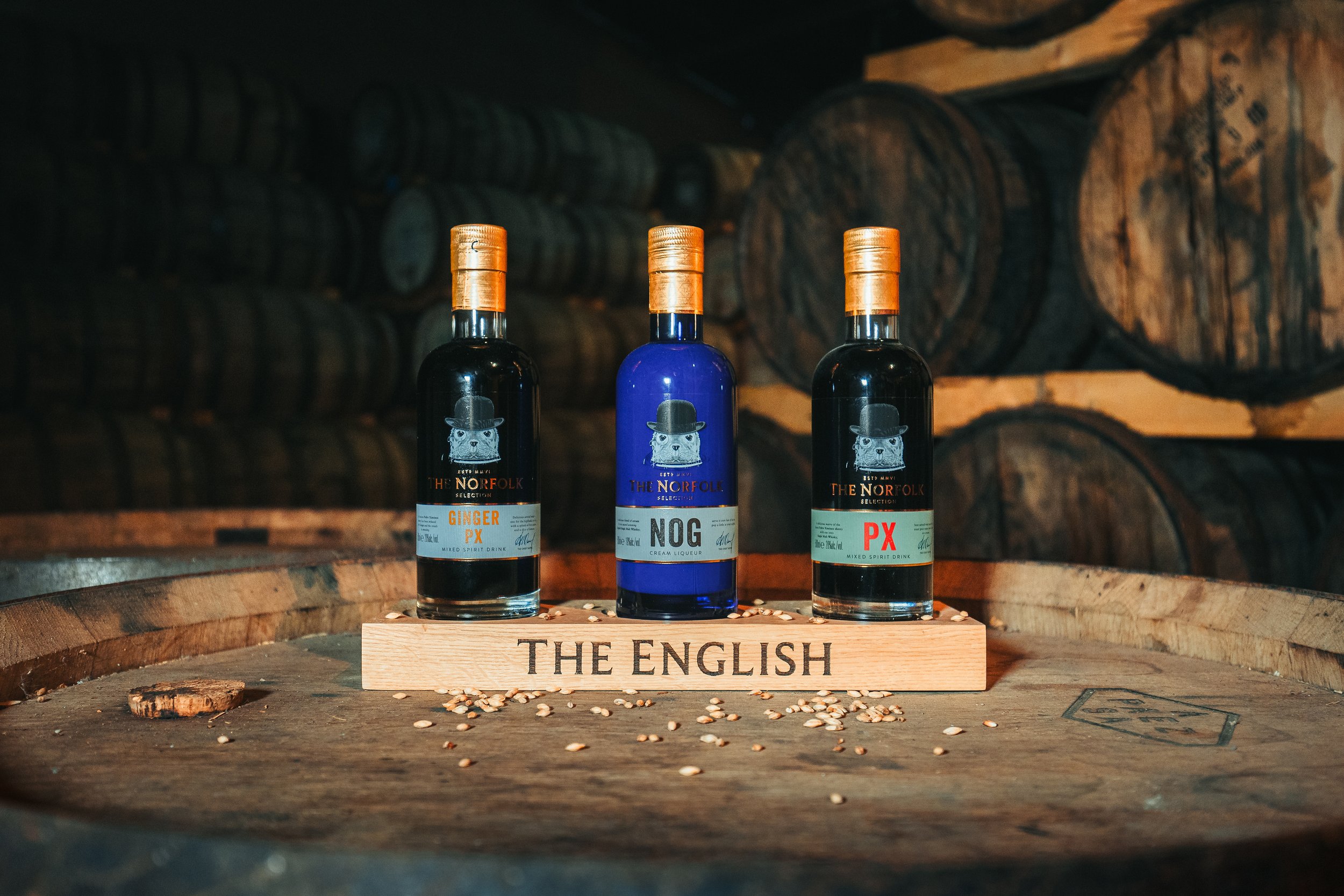 Three bottles of flavored spirit drinks placed on a wooden rack labeled 'The English' on a barrel in a dimly lit cellar with large wooden barrels in the background.