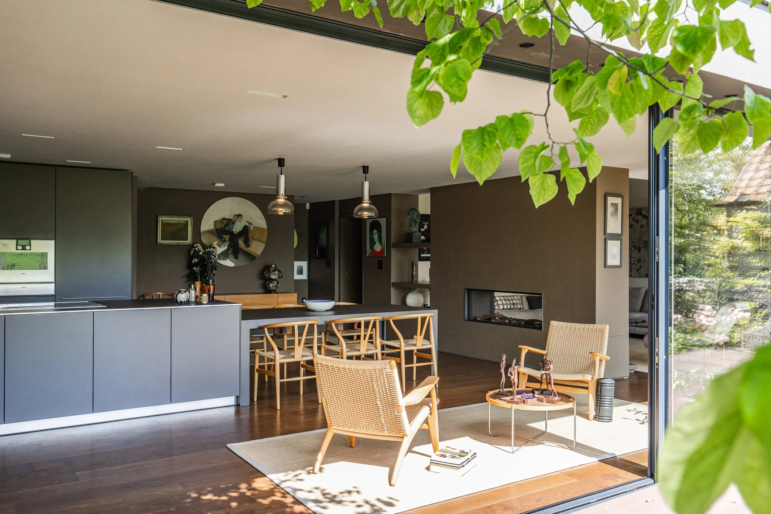Modern open-plan living room with a view into the kitchen, featuring wooden furniture, a white rug, a beige armchair, a coffee table with decorative items, and large glass windows with greenery outside.