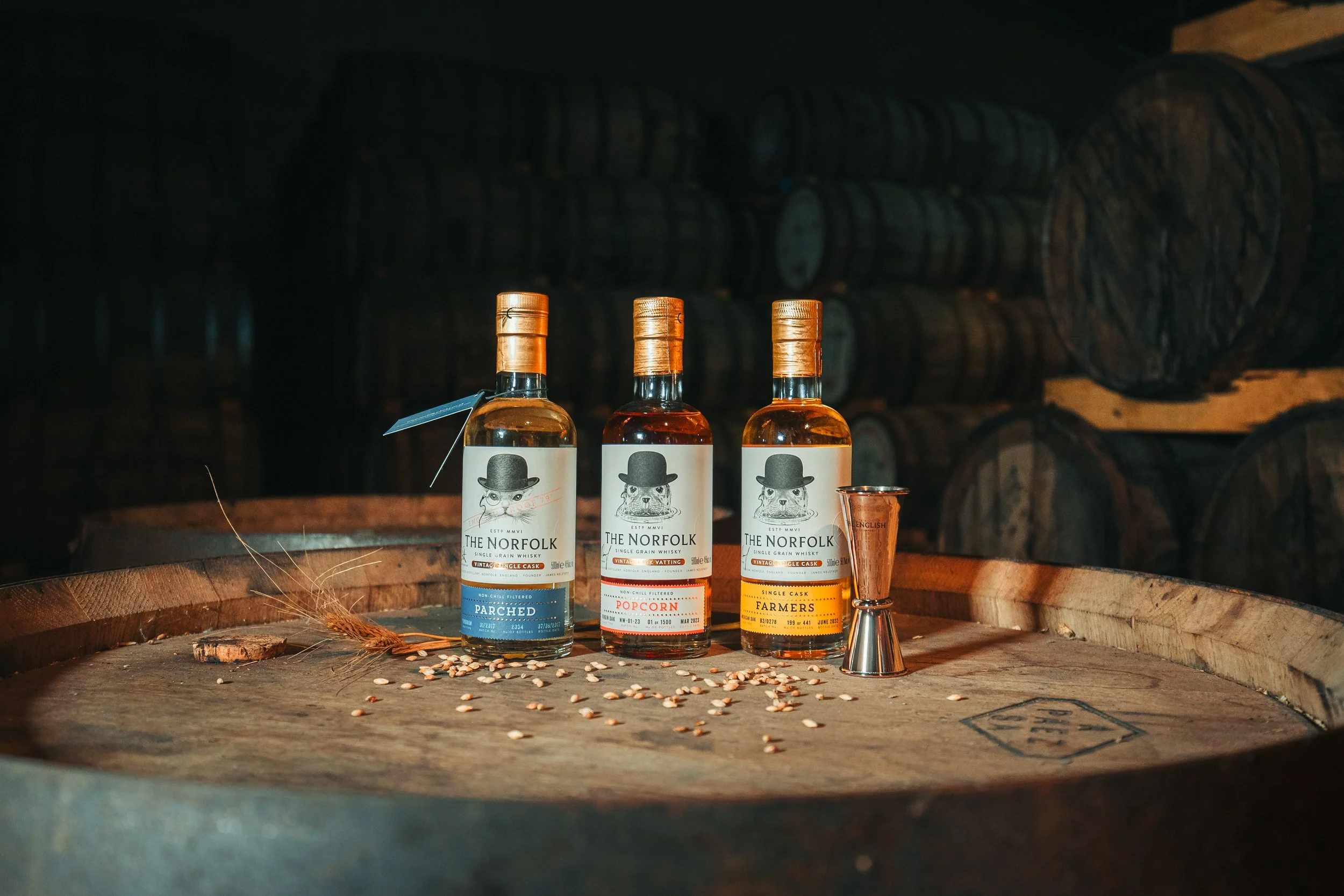 Three bottles of whiskey labeled 'The Norfolk', placed on a wooden barrel in a cellar with barrels stacked in the background.