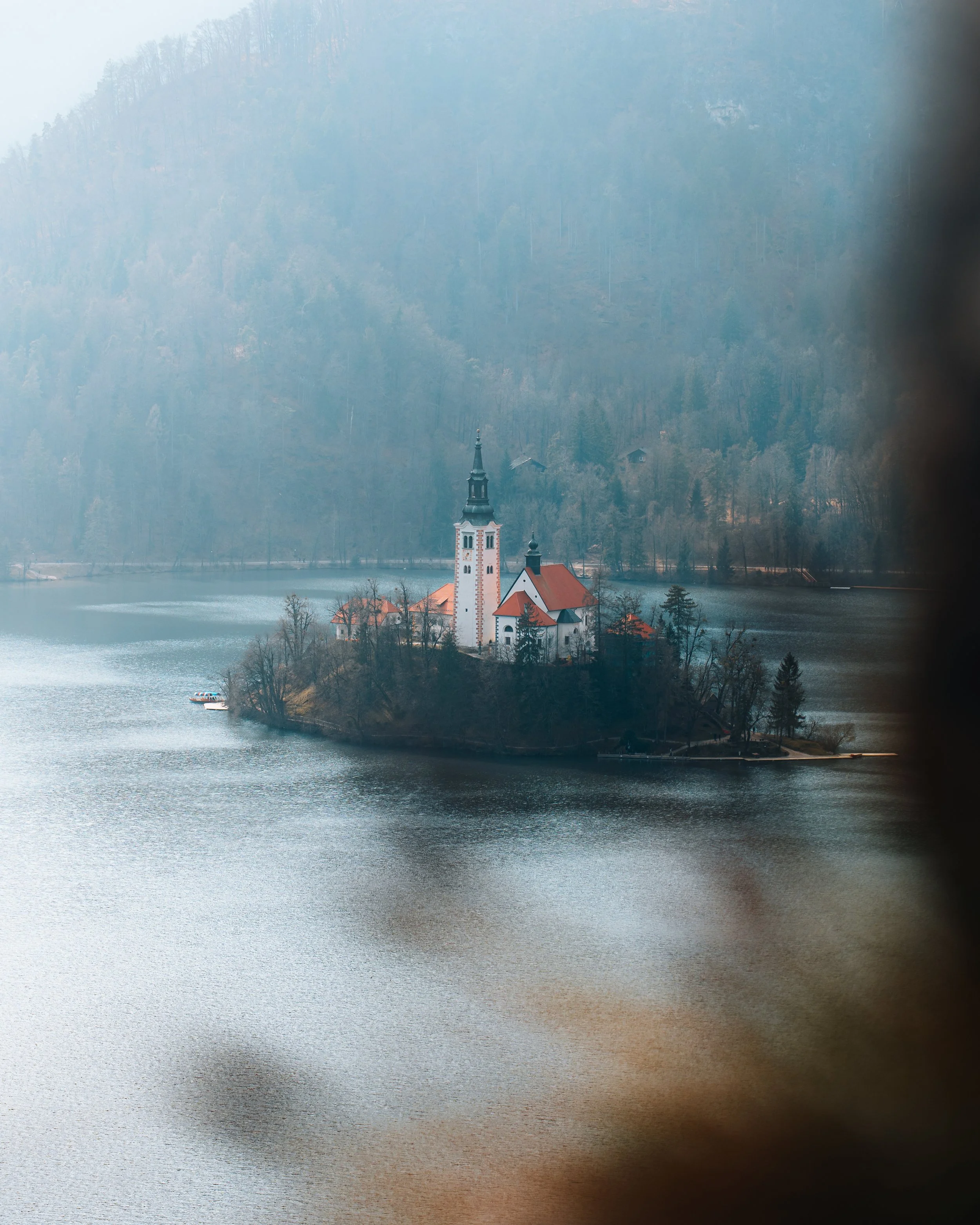 A scenic view of a small church on an island in a lake, surrounded by mountains and trees, viewed through a window.