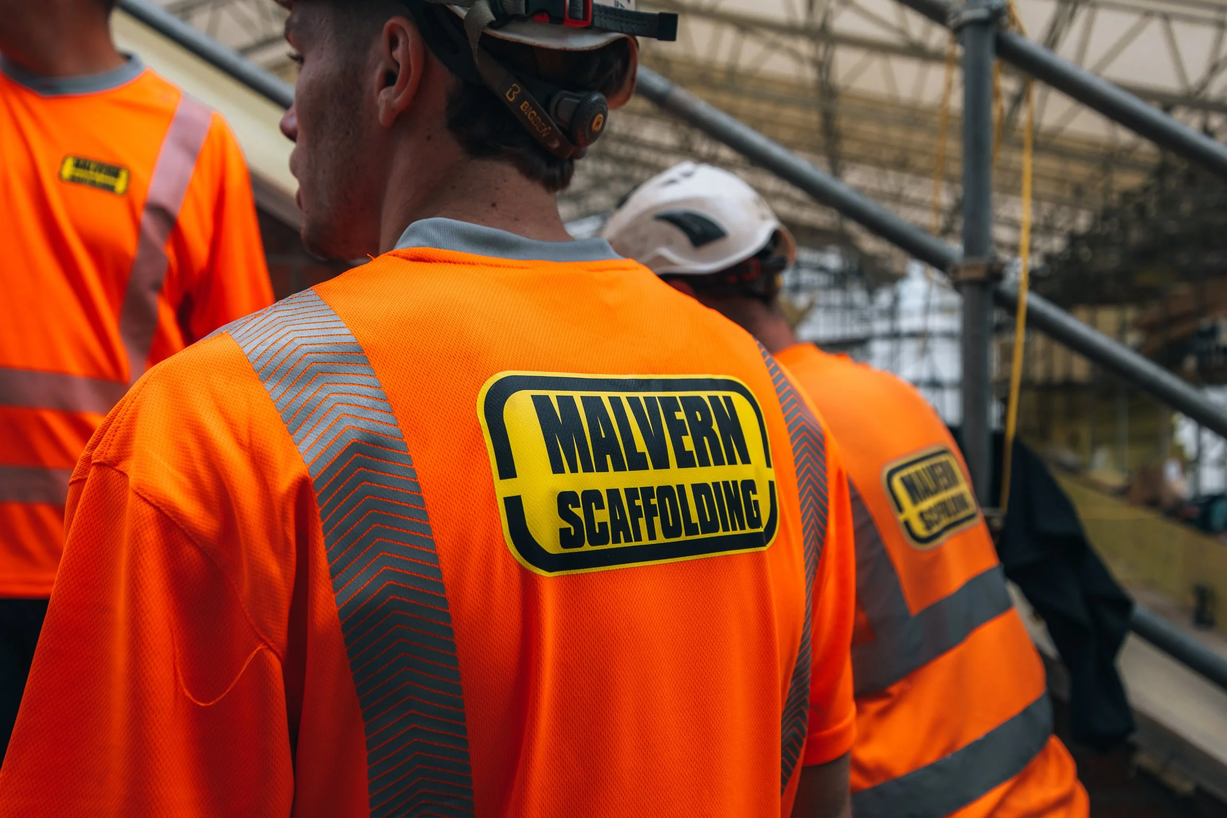 Construction workers wearing bright orange safety vests with the logo 'Malvern Scaffolding,' standing on a scaffolding structure.