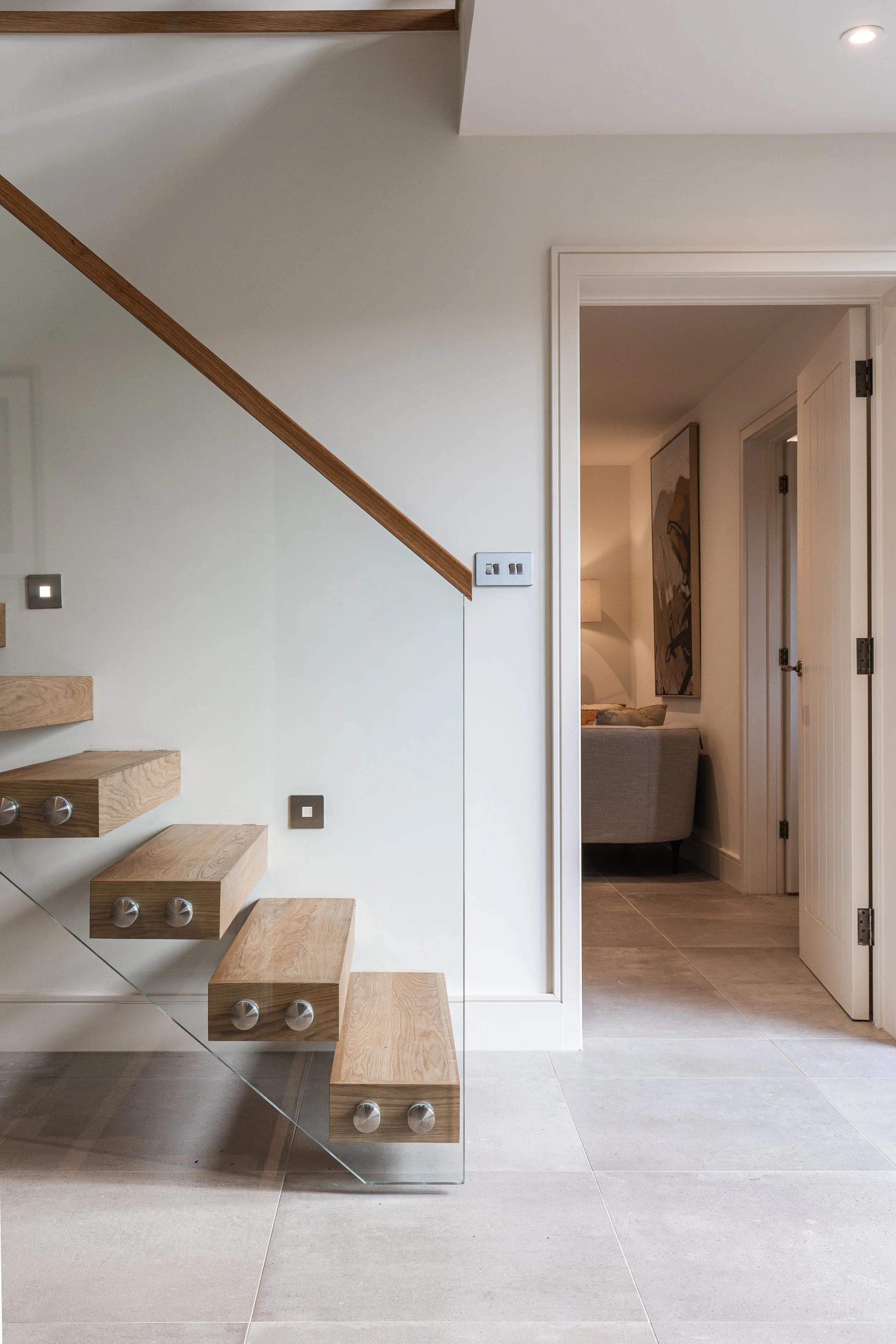 Modern interior space featuring a floating staircase with wooden steps and glass railing next to a white wall and a doorway leading to a living room with a sofa and framed artwork.