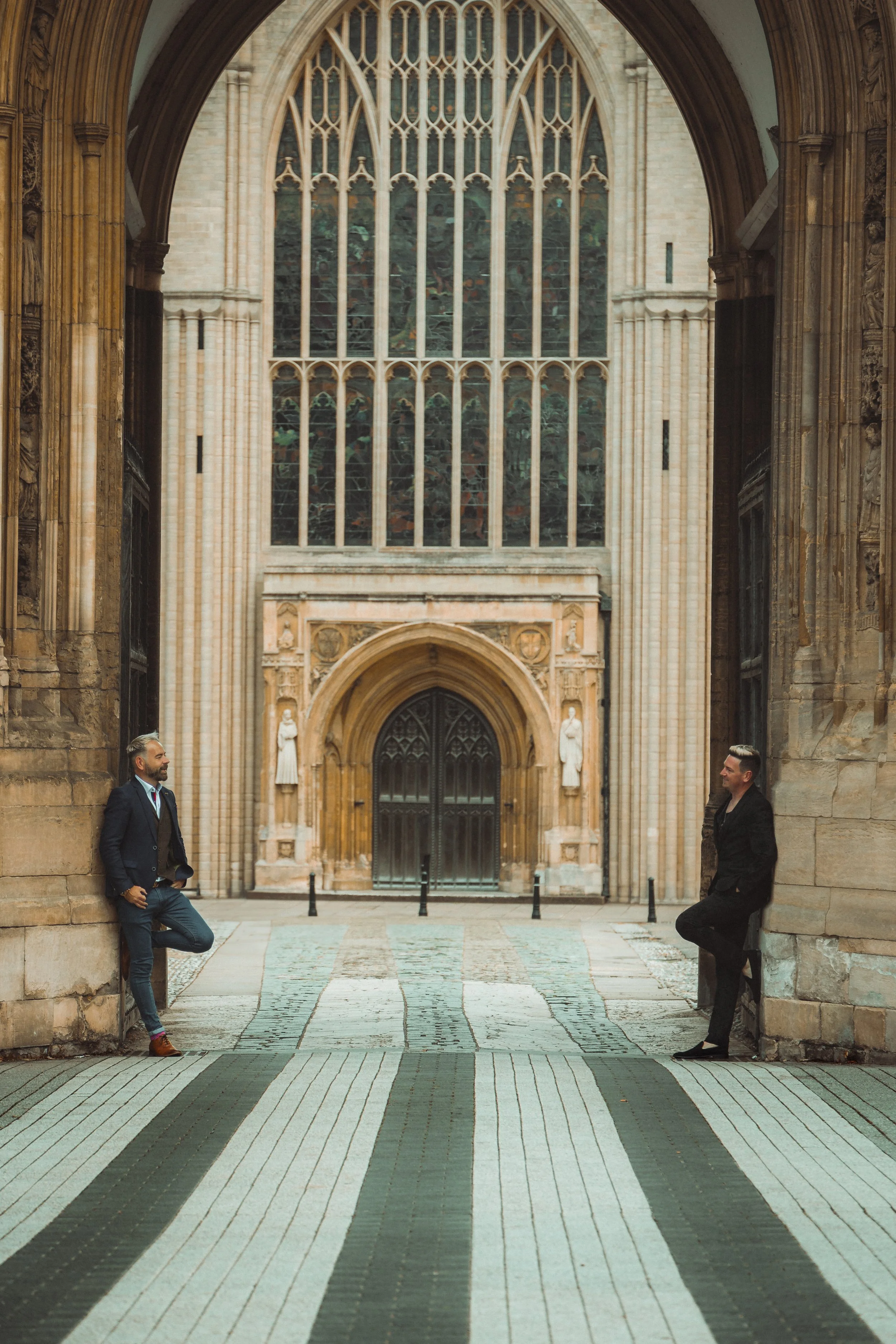 Two men in suits are standing and leaning against the stone walls in a historic, Gothic-style building with large stained glass windows and ornate architecture.