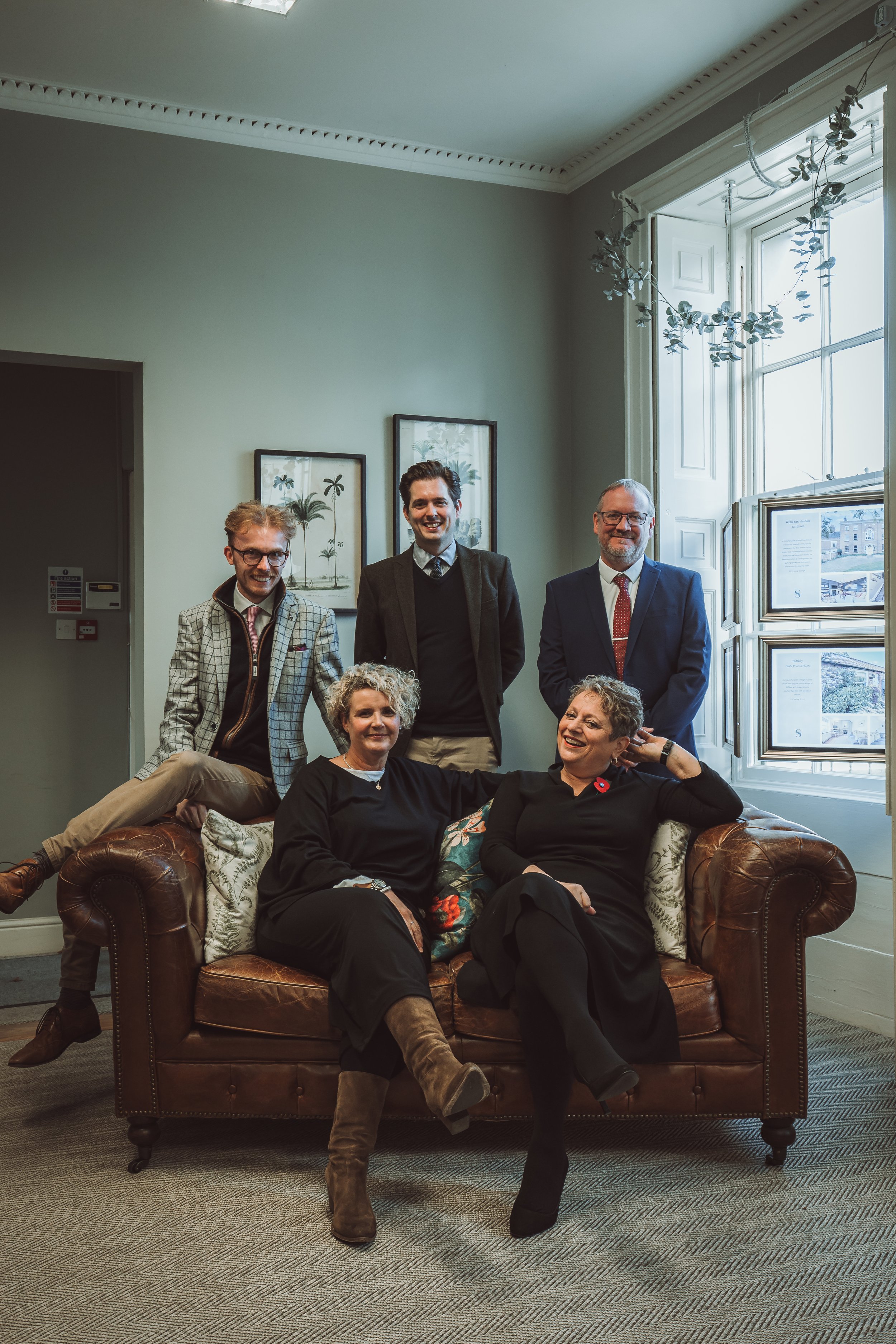Six people posing in a living room, with two women sitting on a leather couch and four men standing behind them. The room has high ceilings, crown molding, and a large window with decor hanging from it.