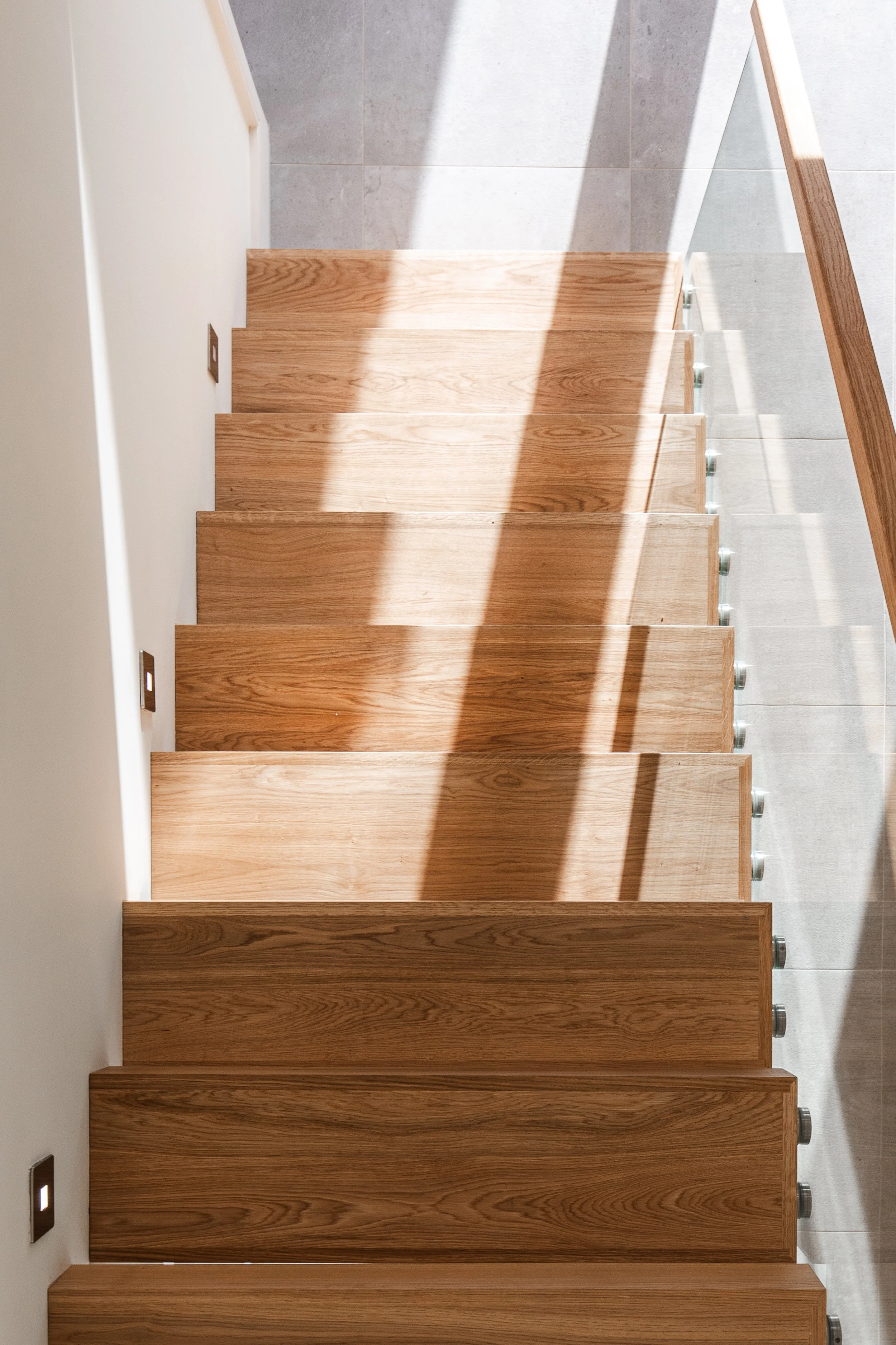 View of wooden staircase with shadow cast on steps, with gray tiled floor and white walls.
