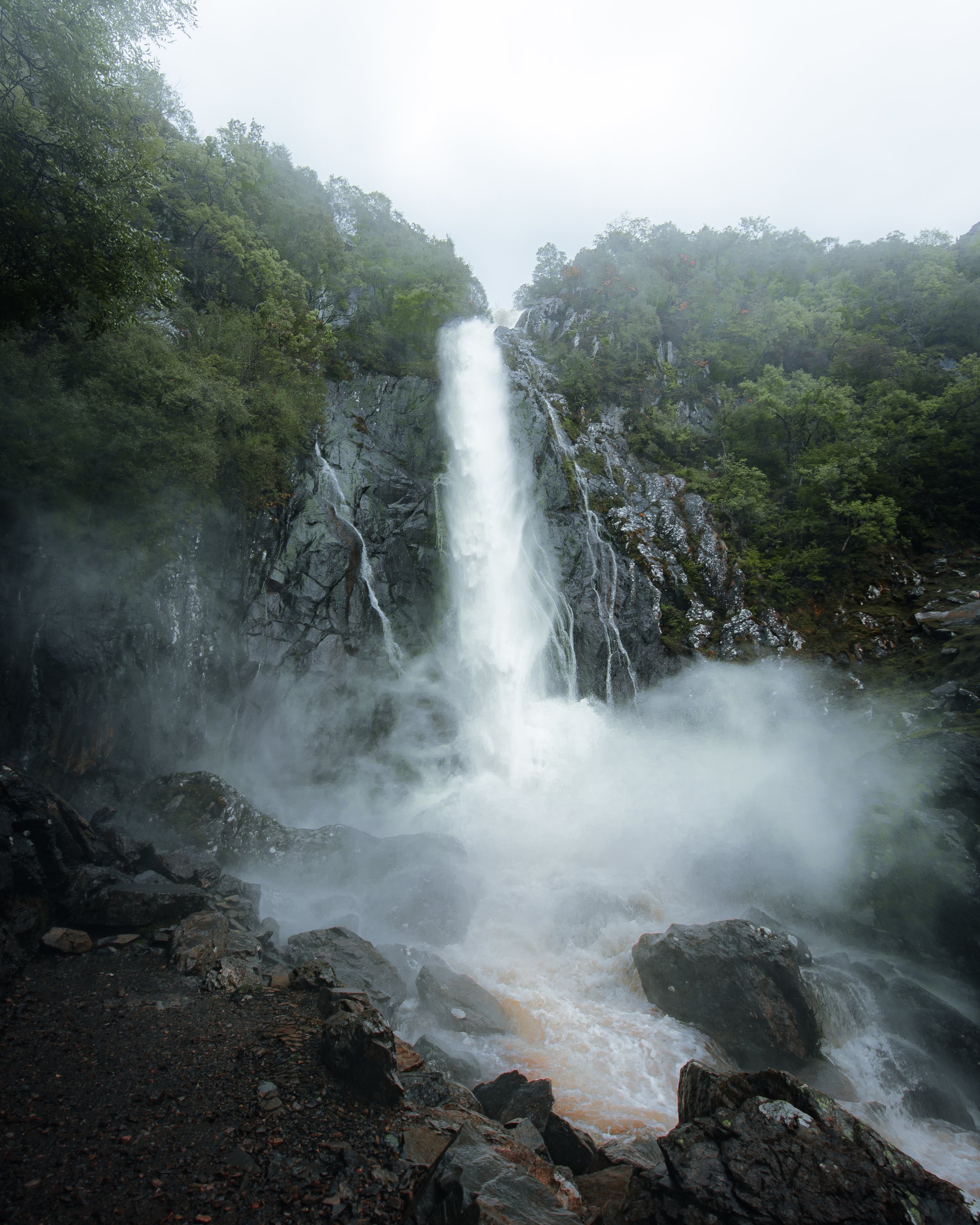 A tall waterfall flowing down a rocky cliff surrounded by green trees and fog.