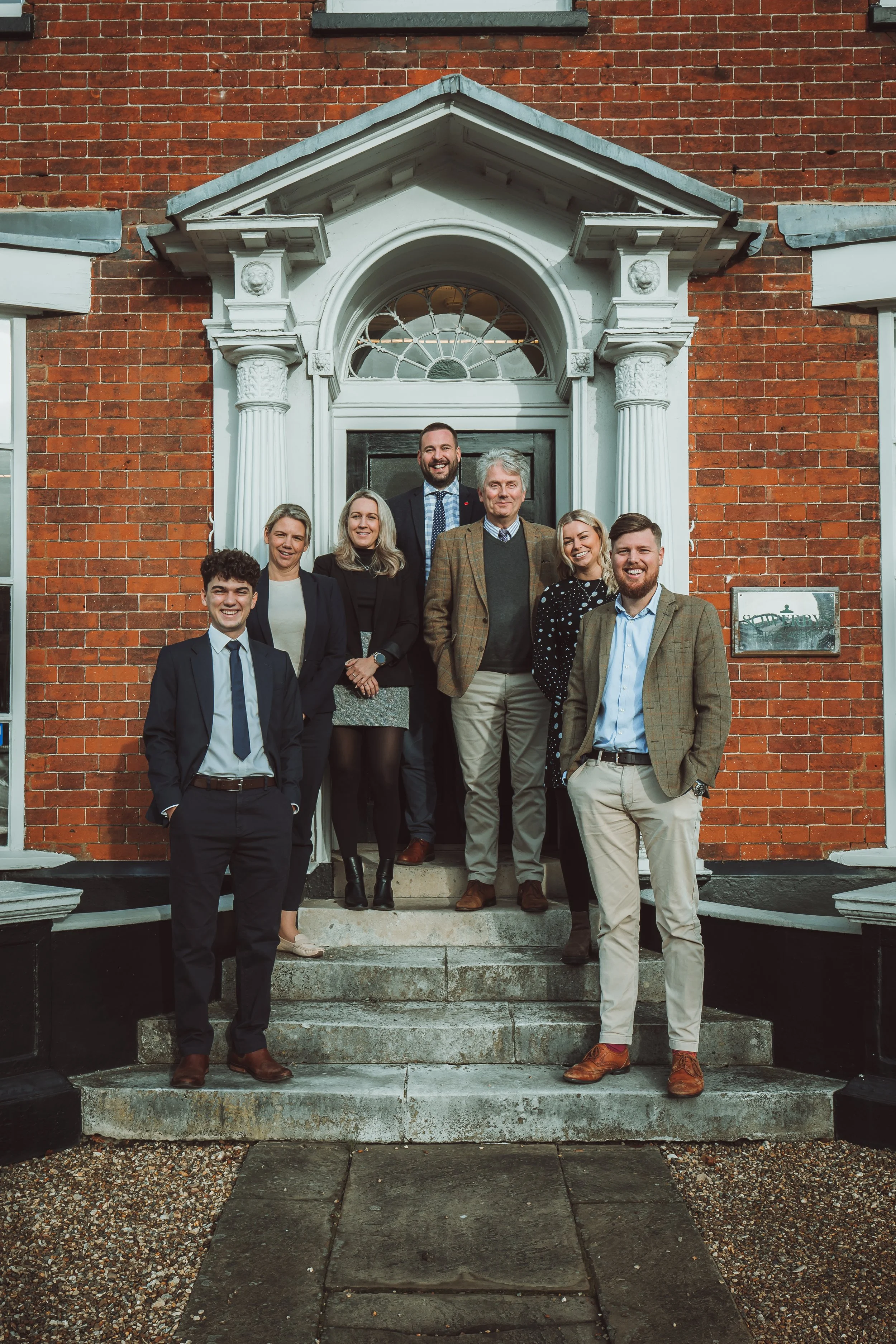 A group of seven professionally dressed people standing on the steps of a brick building with a decorative white doorframe and a black door, smiling at the camera.
