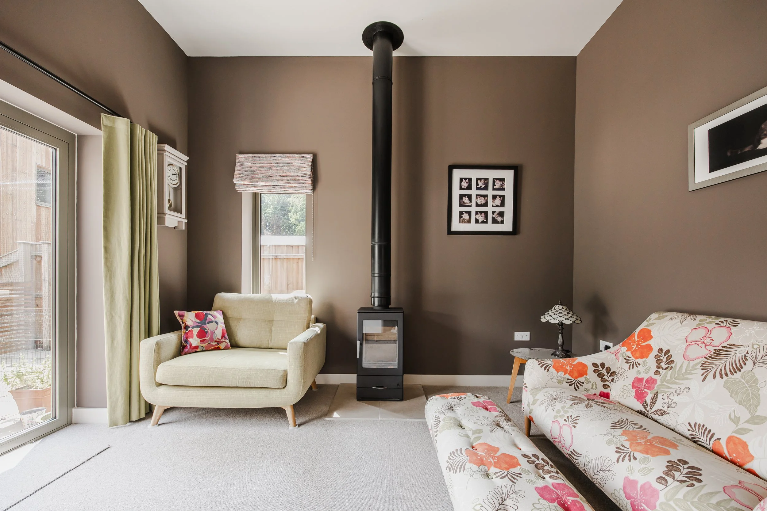 Living room with beige armchair and floral patterned sofa, small side table with lamp, wood stove against a brown wall, framed photos, window with patterned Roman blind, and sliding glass door with green curtains.