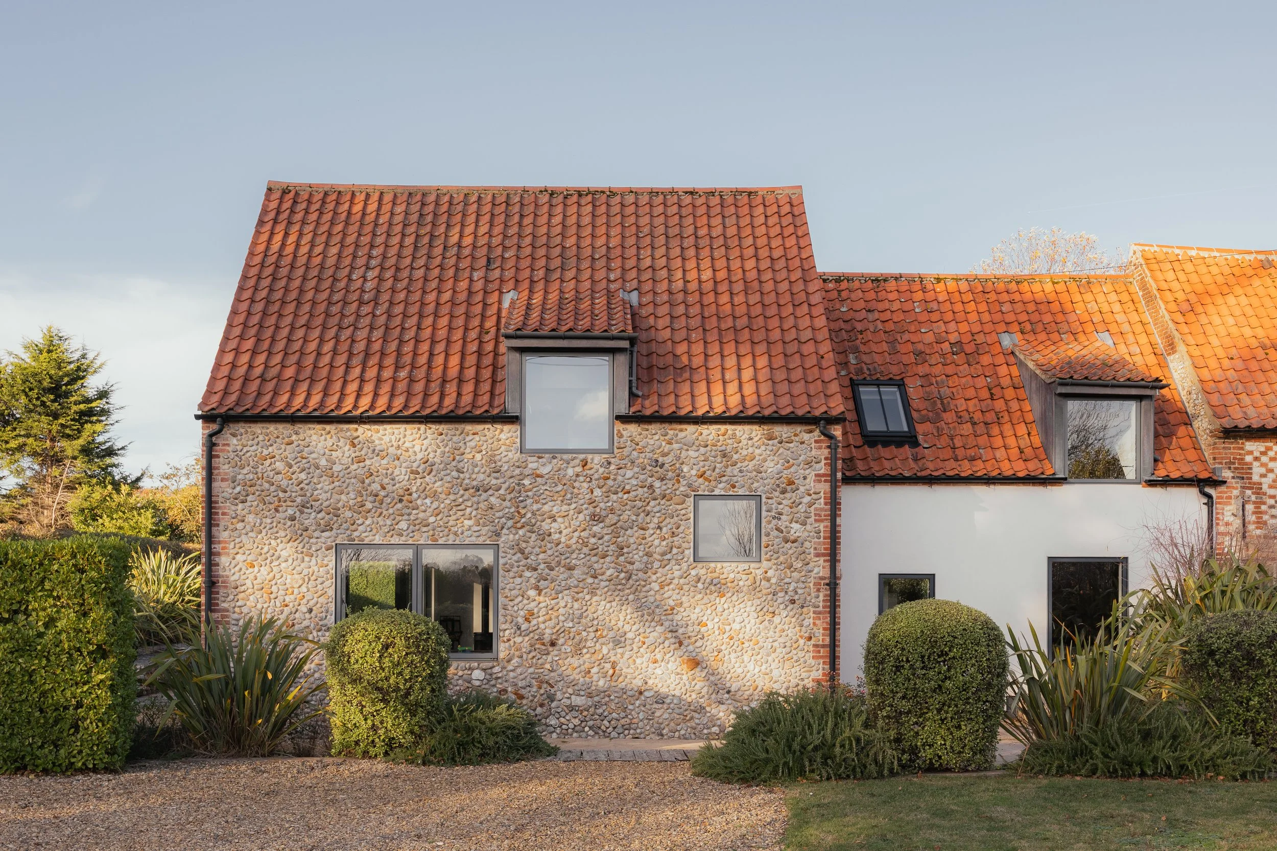 A house with a red-tiled roof, stone and white exterior walls, and various windows, surrounded by shrubbery and plants, during daytime.