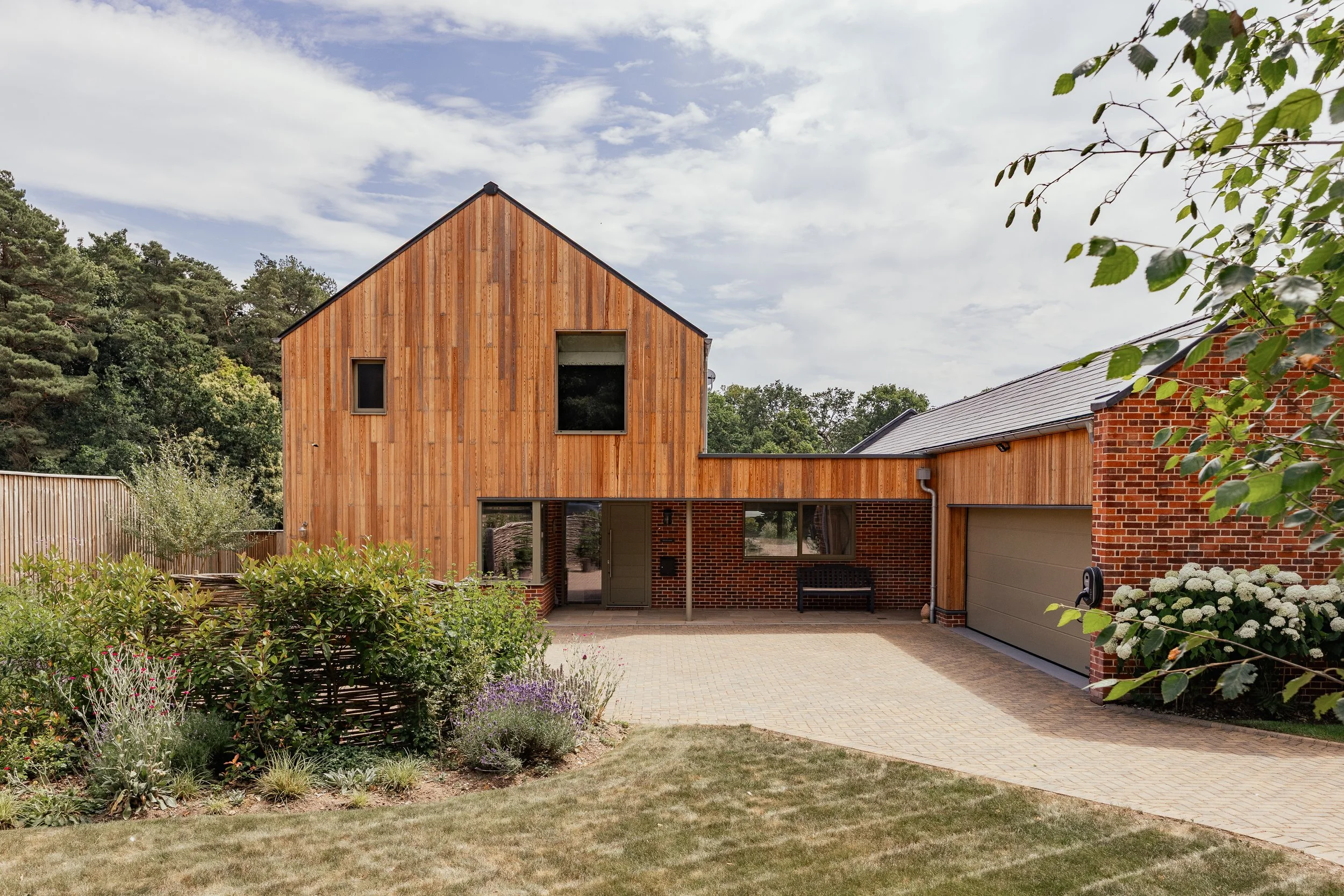 A modern house with a wooden facade, brick accents, and a gray garage door, surrounded by a landscaped yard with bushes, flowers, and a lawn, under a partly cloudy sky.