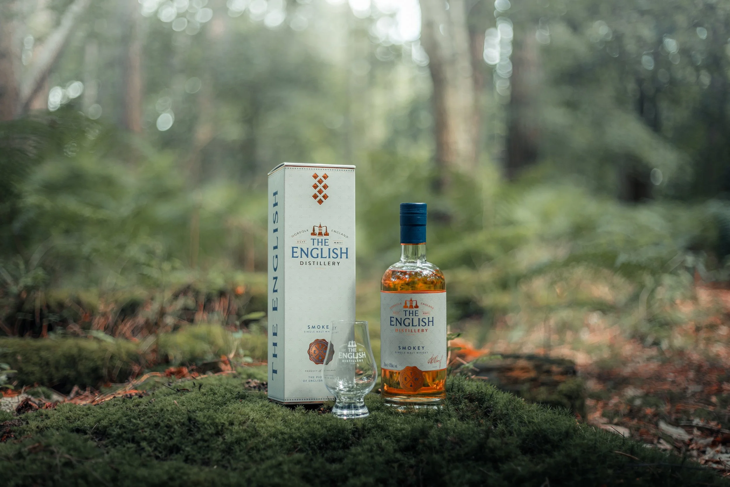 A bottle of The English Distillery Smokey single malt whiskey, a branded glass, and the whiskey box are placed on mossy ground in a forest with tall trees in the background.