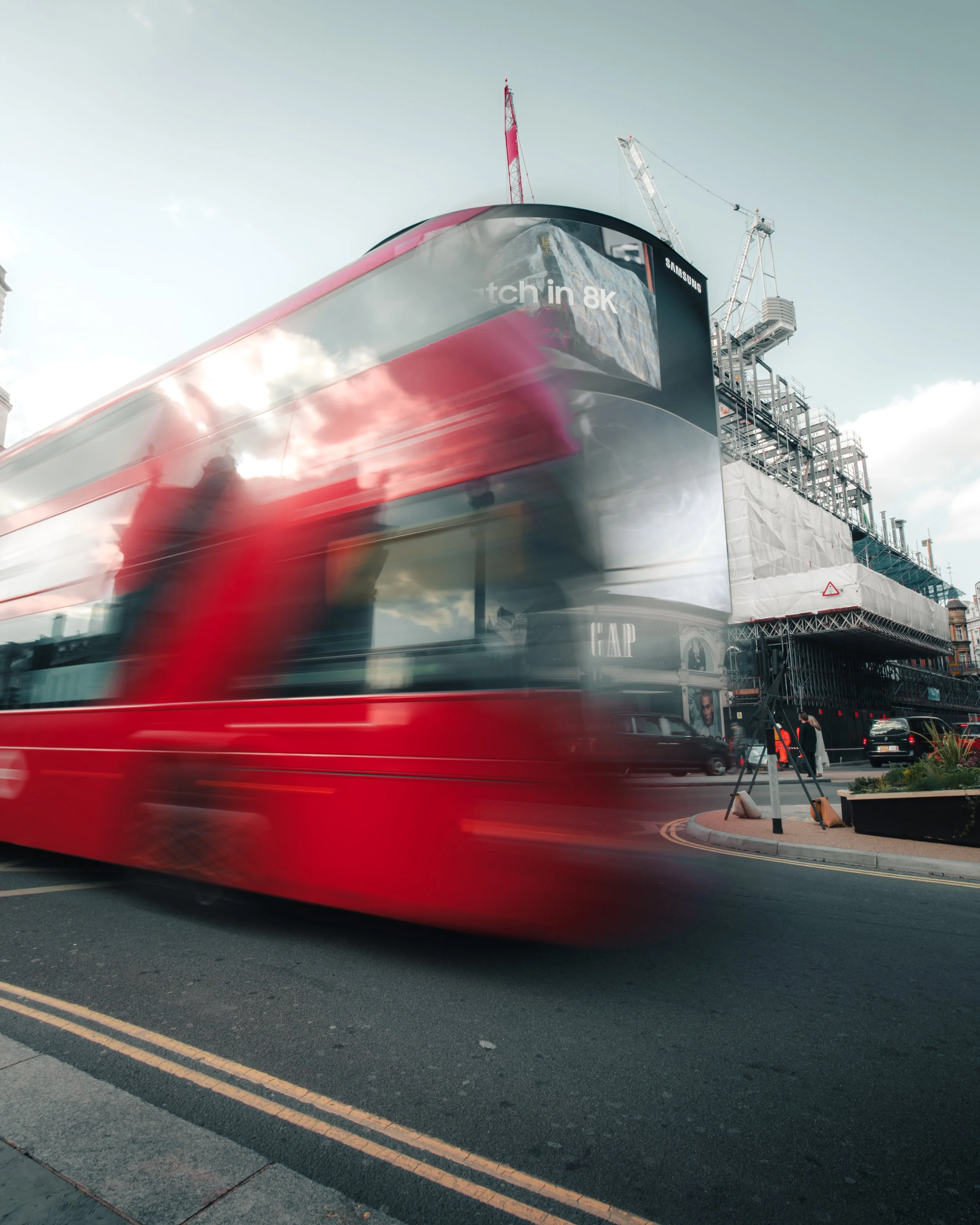 A busy city street with a red double-decker bus in motion, blurred due to speed, passing by skyscrapers under construction with cranes, and advertisements on buildings.