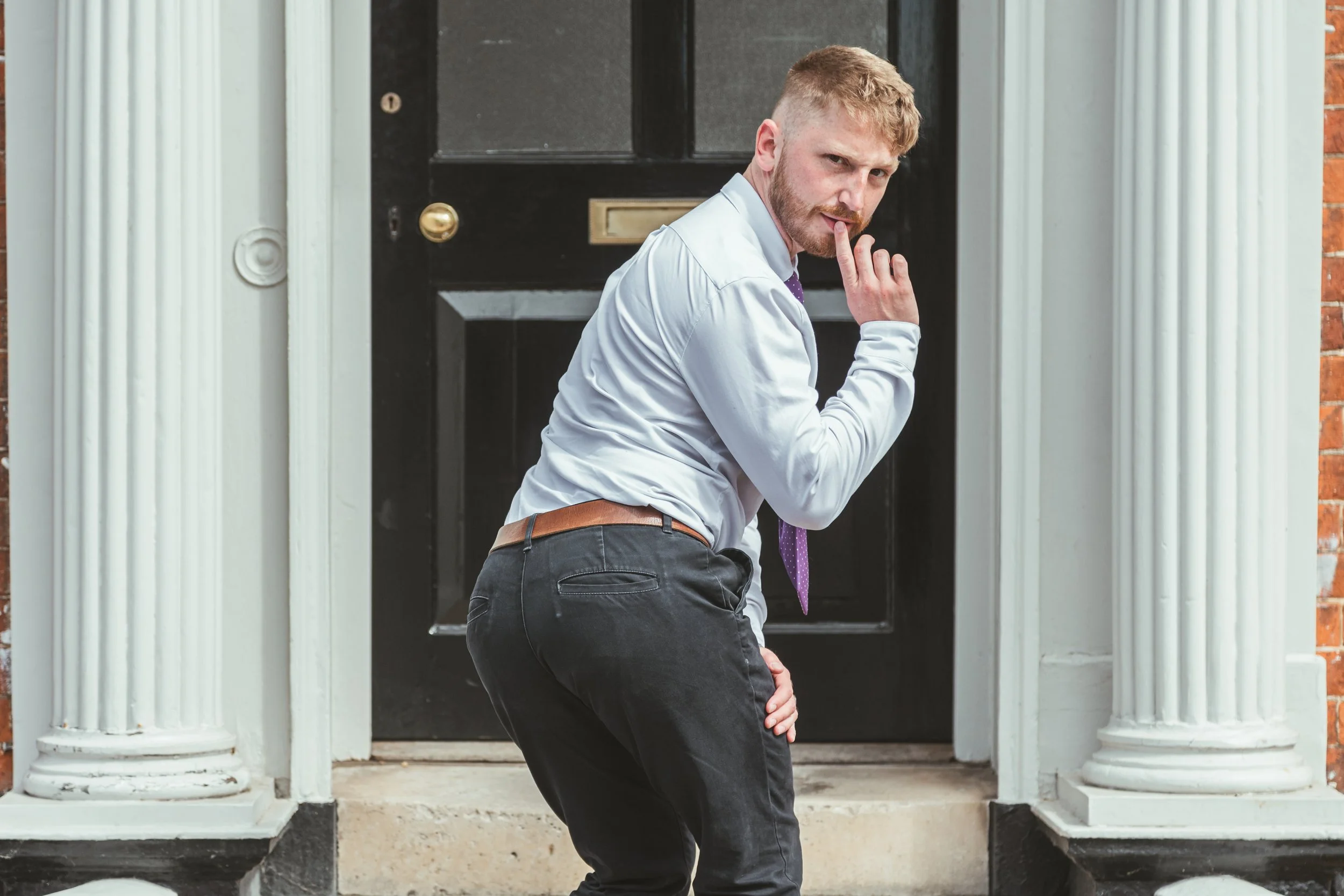 A man with light hair and facial hair, wearing a white dress shirt and black pants, is standing outside a black front door with white columns, making a gesture with his hand near his lips, looking over his shoulder.