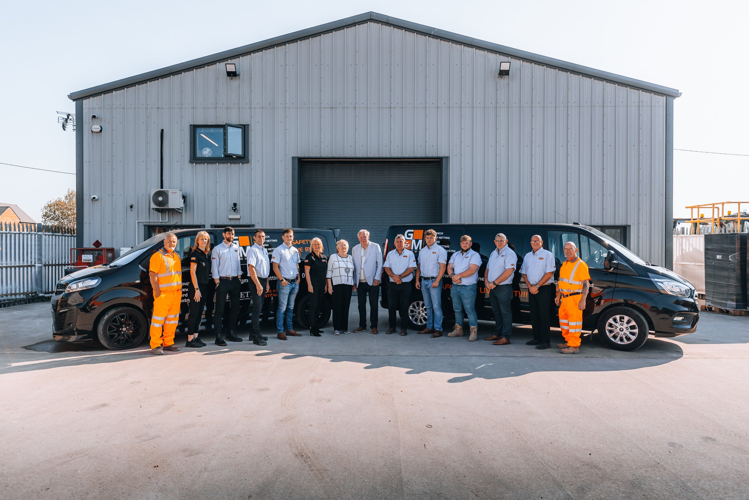 Group of people standing between two black vans in front of a gray industrial building, some wearing white shirts and others in orange safety clothing.