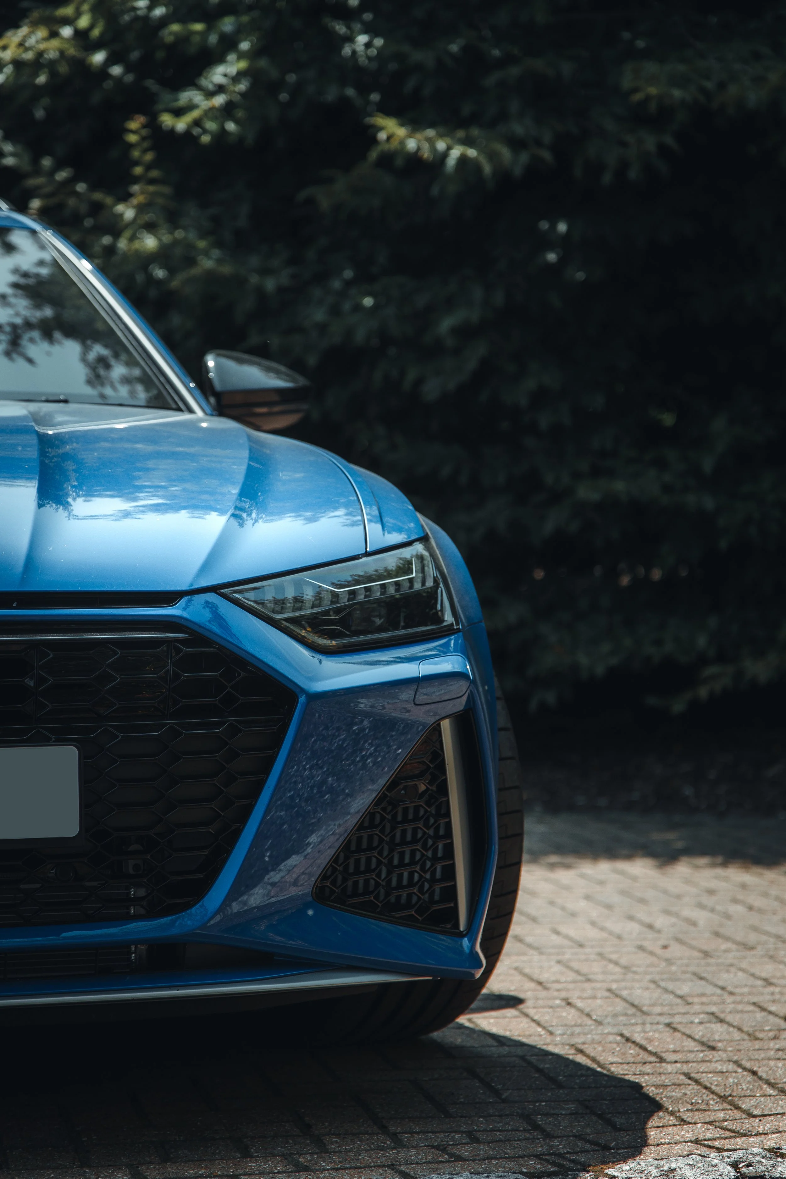 Close-up of the front end of a blue sports car parked on a brick surface, with a black honeycomb grille, angular headlights, and a background of green trees.