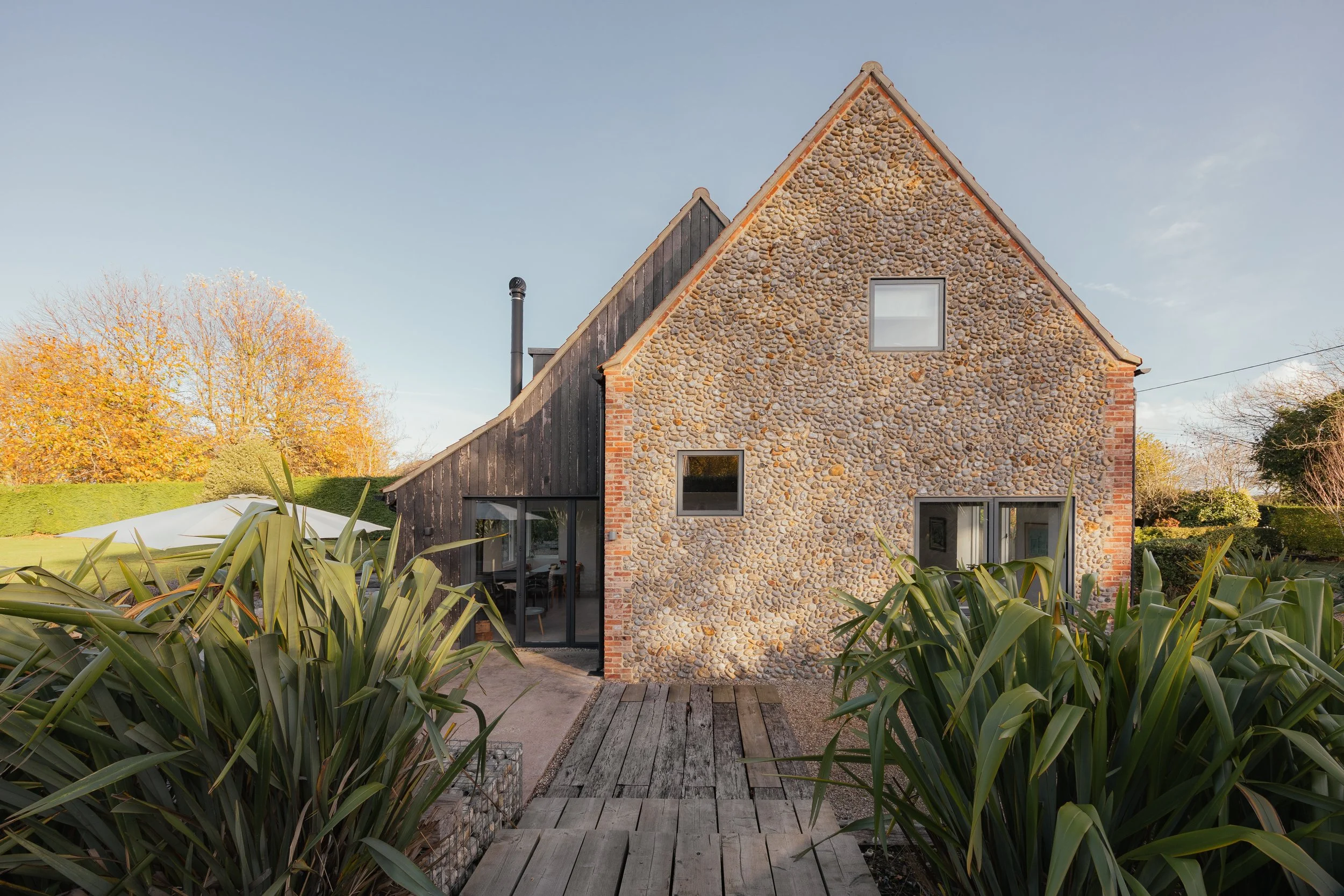 Modern house with pebble stone façade and large glass doors, surrounded by green plants and trees with autumn leaves, under a clear blue sky.