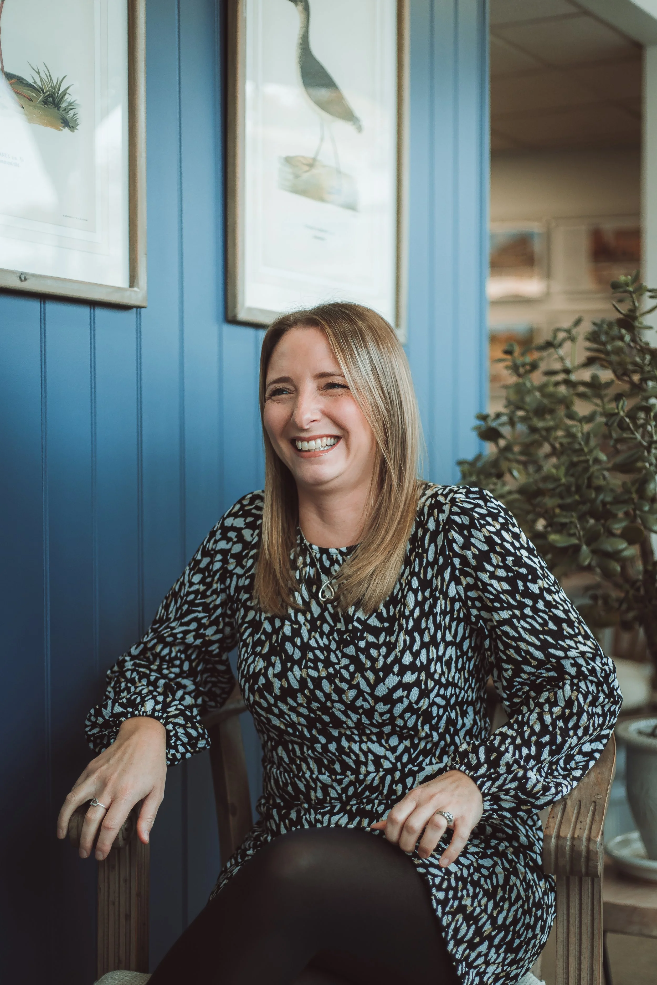 A smiling woman with shoulder-length blonde hair, wearing a black and white patterned dress, is seated indoors against a blue paneled wall decorated with framed artwork, with plants nearby.