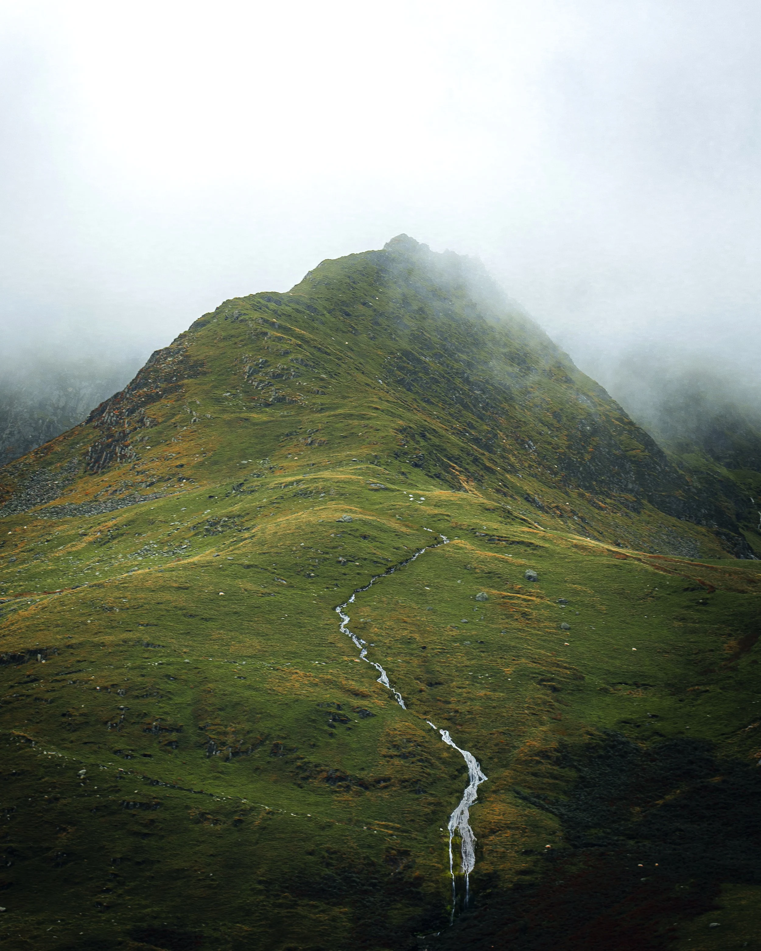 A green mountain with a small stream flowing down its slope, partially covered by fog.