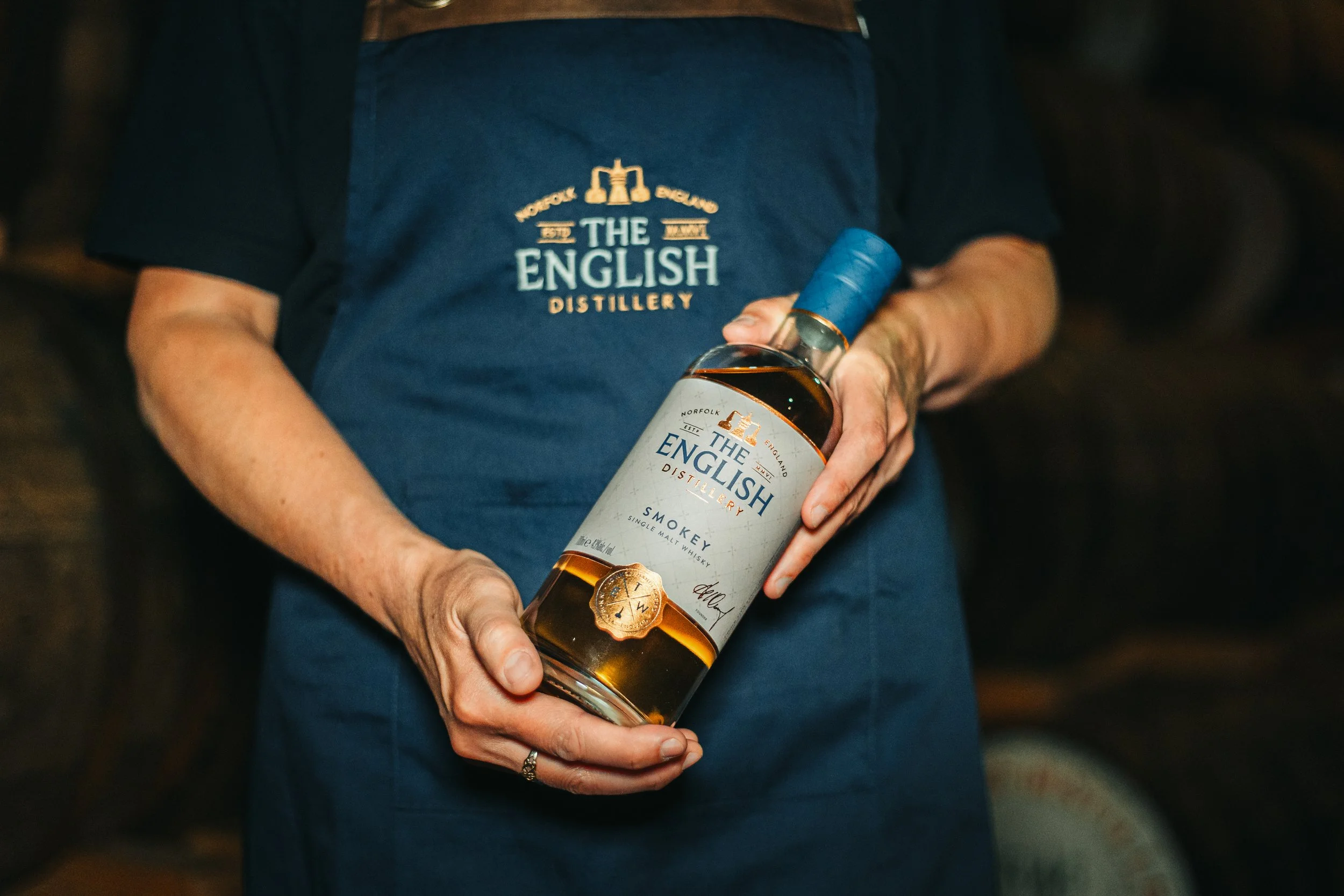 Person wearing a blue apron with 'The English Distillery' logo holding a bottle of Smokey single malt whiskey.