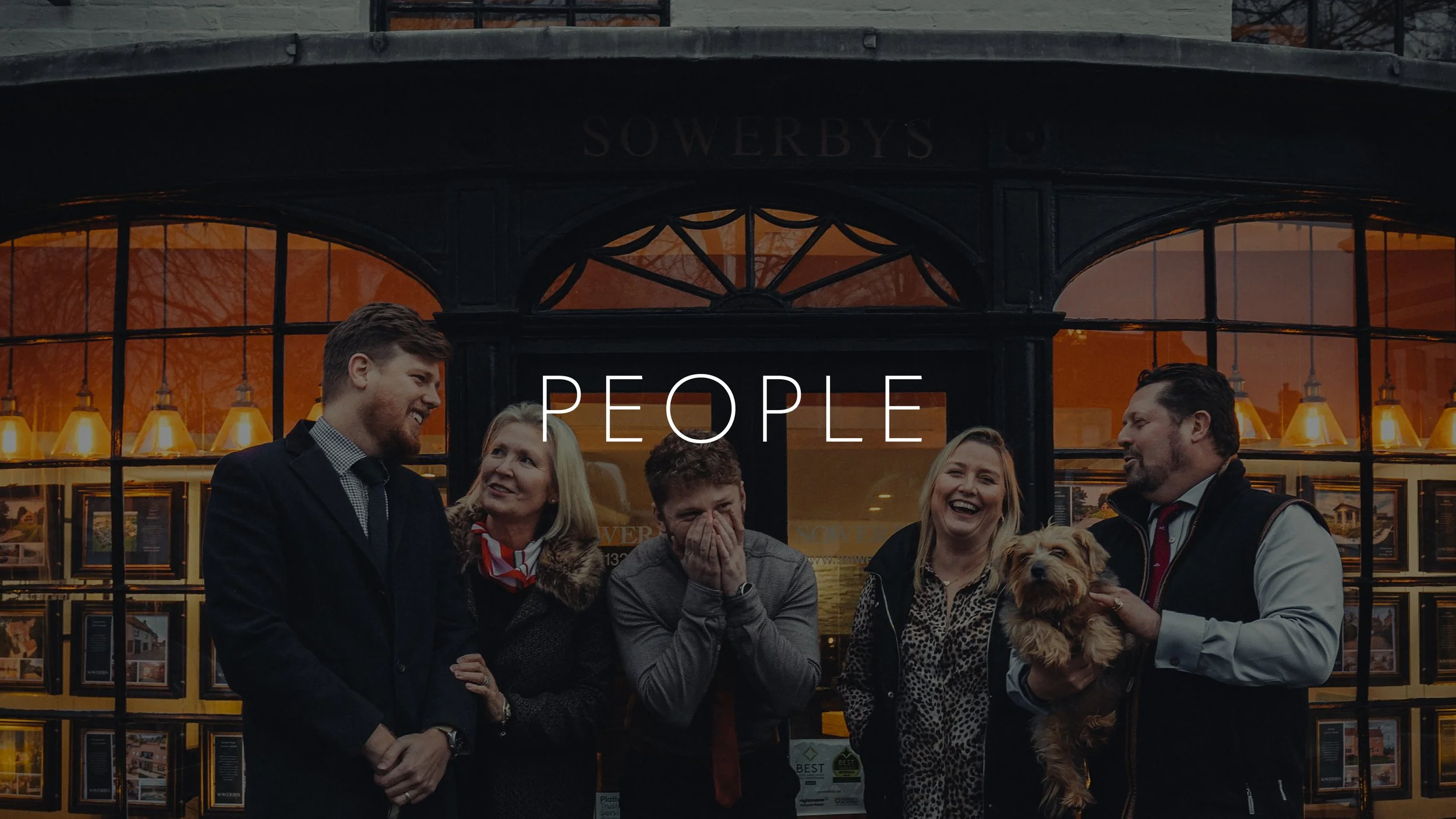 Group of five people standing in front of a building with the word 'Sowerbys' on it, socializing and smiling, with one person holding a small dog.