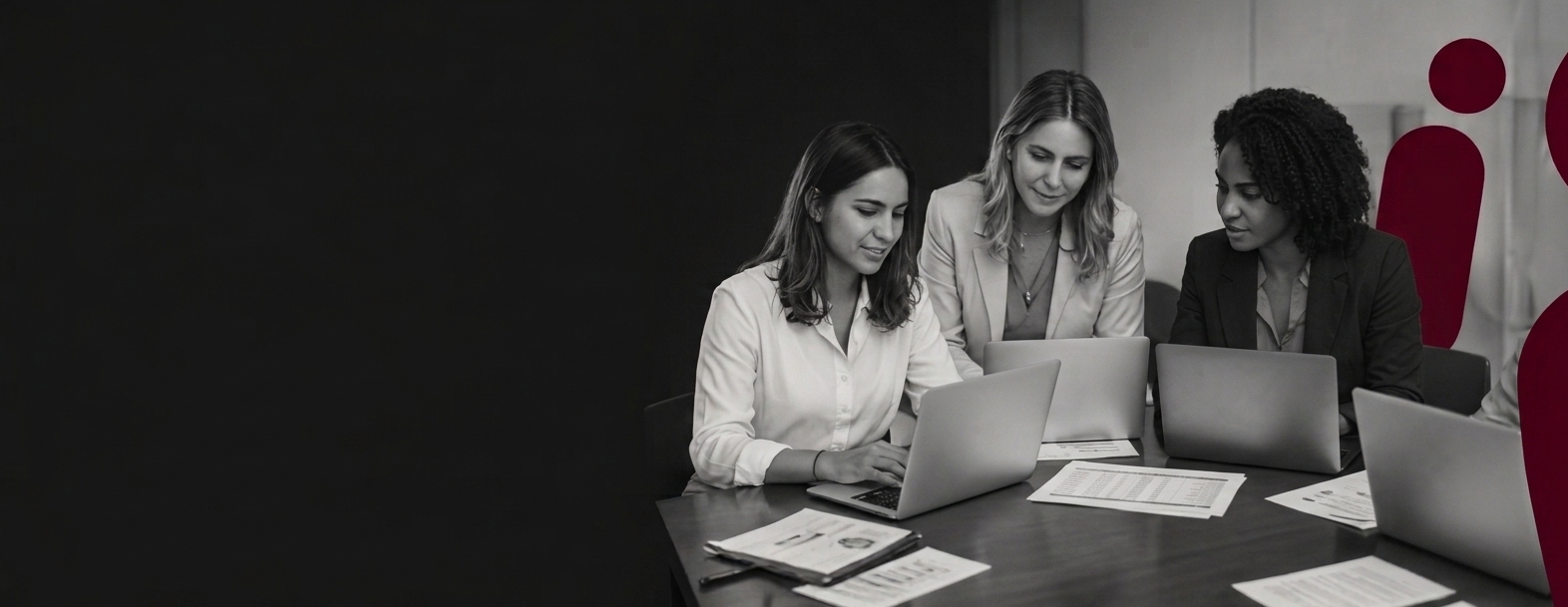 Three women working together at a conference table with laptops and documents.