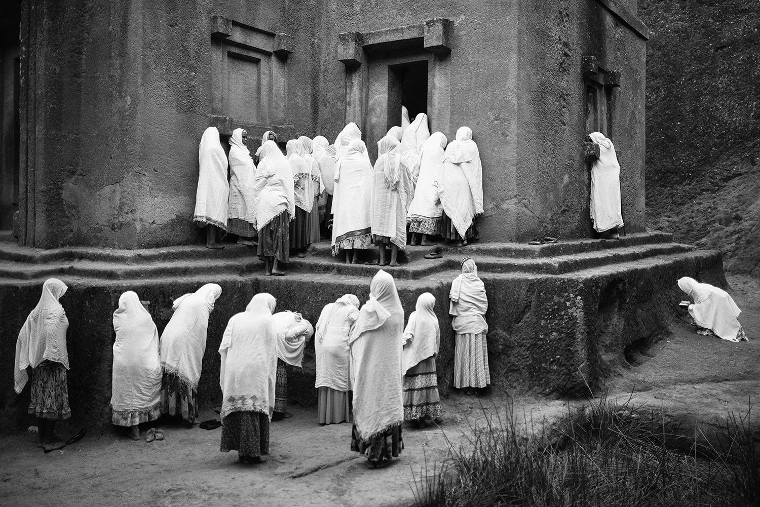 Grupo de personas viste en ropa tradicional, algunas de ellas esperando en las escaleras y otras en el suelo frente a una estructura de adobe, probablemente en un contexto religioso o cultural.