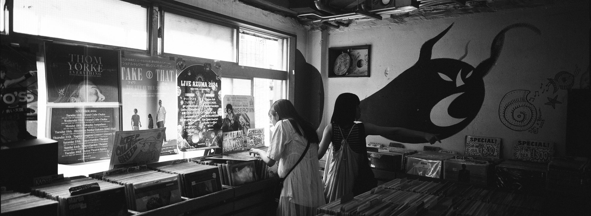 Dos mujeres en una tienda de discos mirando vinilos, con cartelera de eventos y una pared decorada con un mural de un carnero.