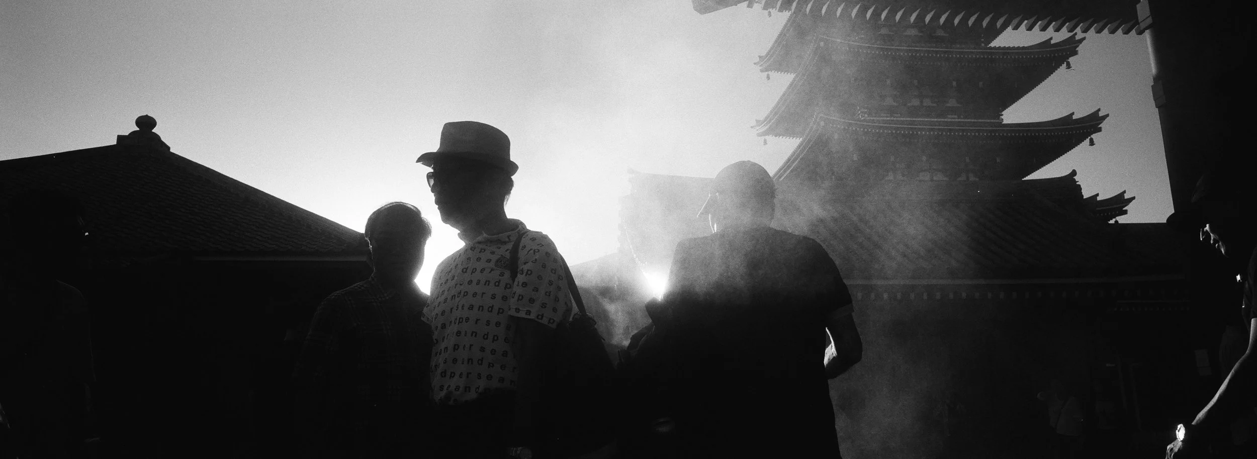Personas caminando frente a un templo pagoda al atardecer, en silueta en una imagen en blanco y negro.
