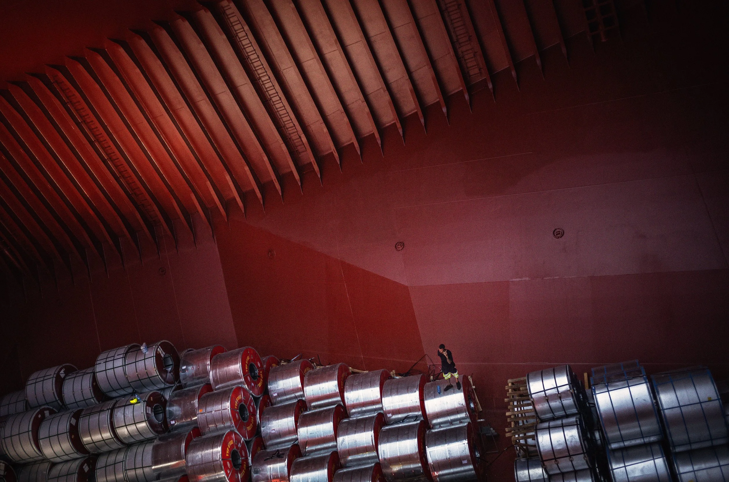 Vista desde arriba del interior de un edificio con una pared de color rojo y varios cilindros metálicos apilados en el suelo, además de una persona caminando entre ellos.