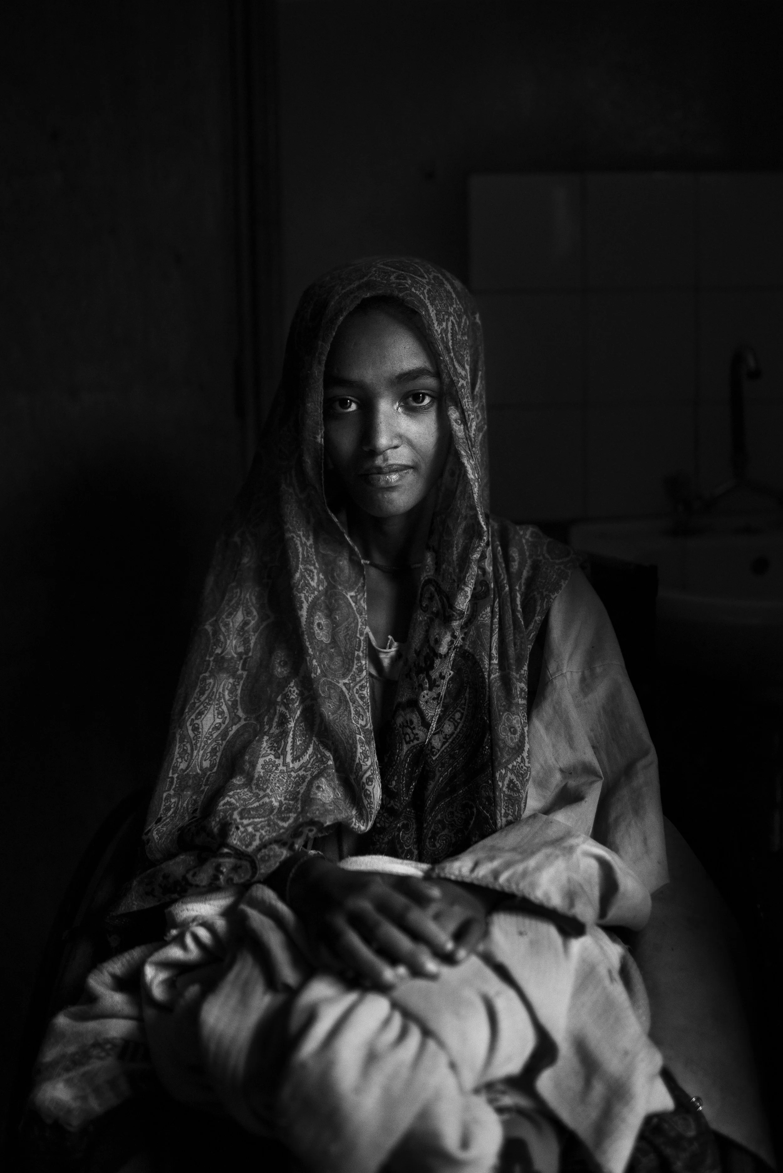 Retrato en blanco y negro de una mujer sentada, mirando hacia la cámara, con un paño en la cabeza y ropa tradicional, en un ambiente con poca luz.