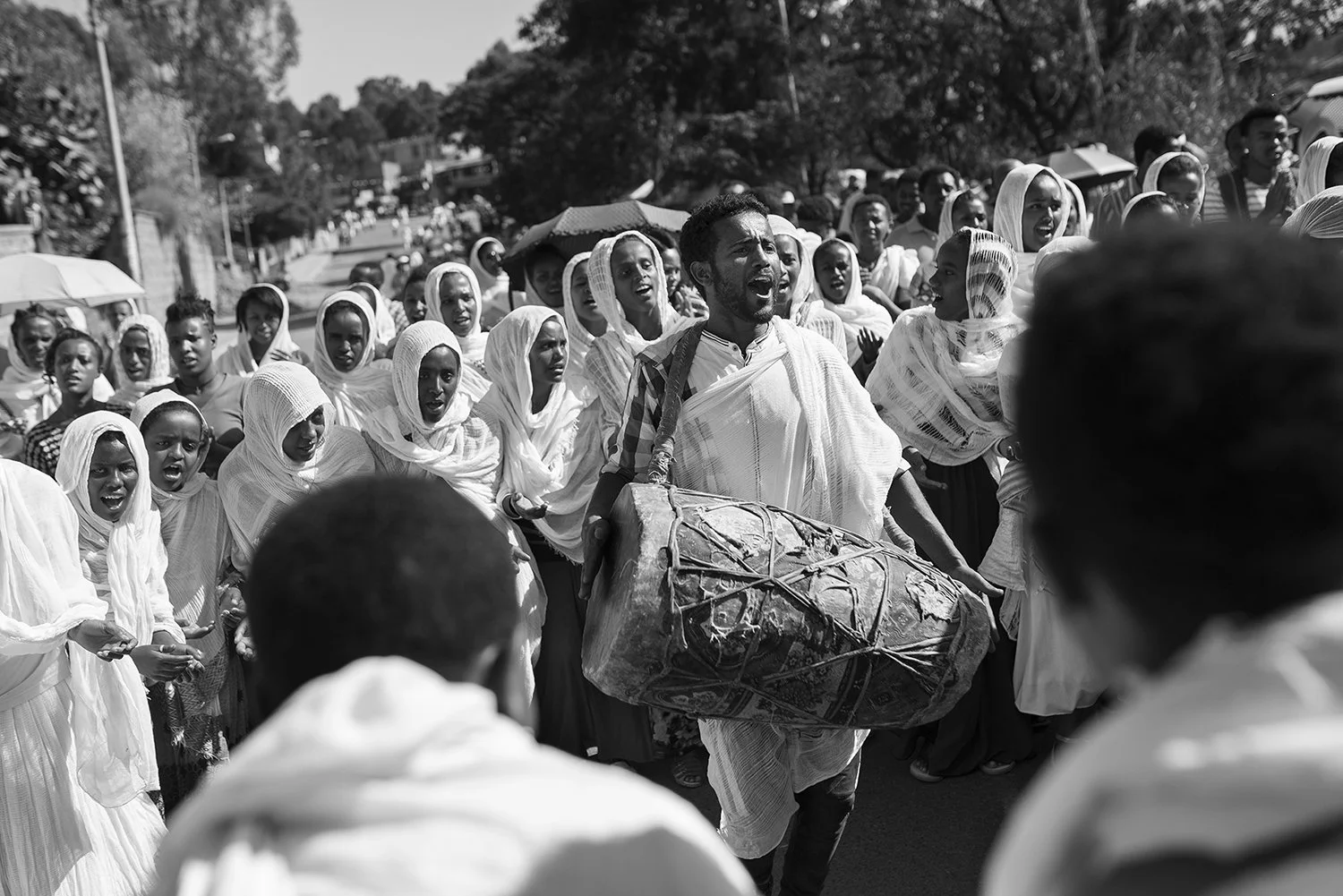 Un grupo de personas con vestimenta tradicional participando en una procesión o danza en la calle, en un día soleado, con árboles y casas de fondo, en negro y blanco.