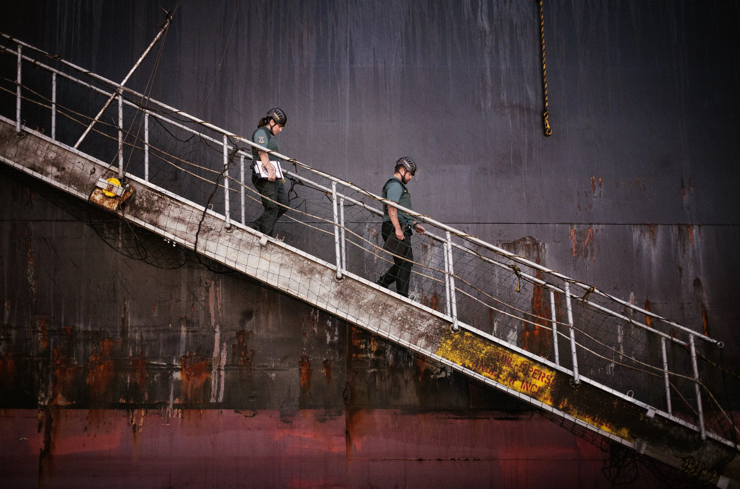 Tres personas con cascos bajando por una escalinata en la parte exterior de un barco oxidado.