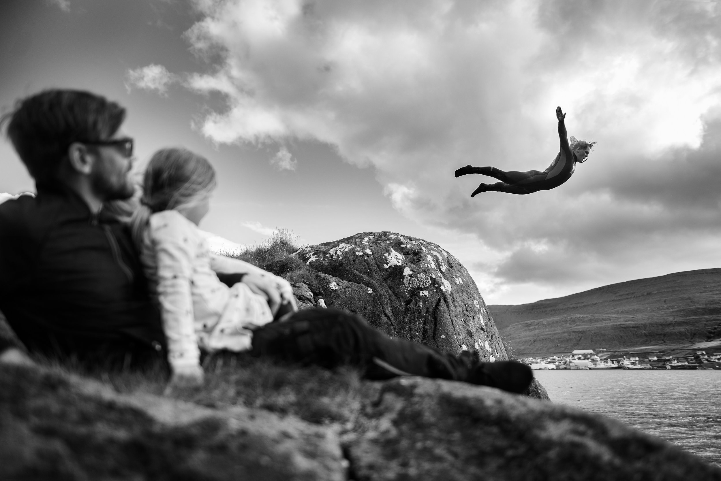 Una pareja y una niña observan a una mujer saltando desde una roca al agua en un paisaje de montaña, en blanco y negro.