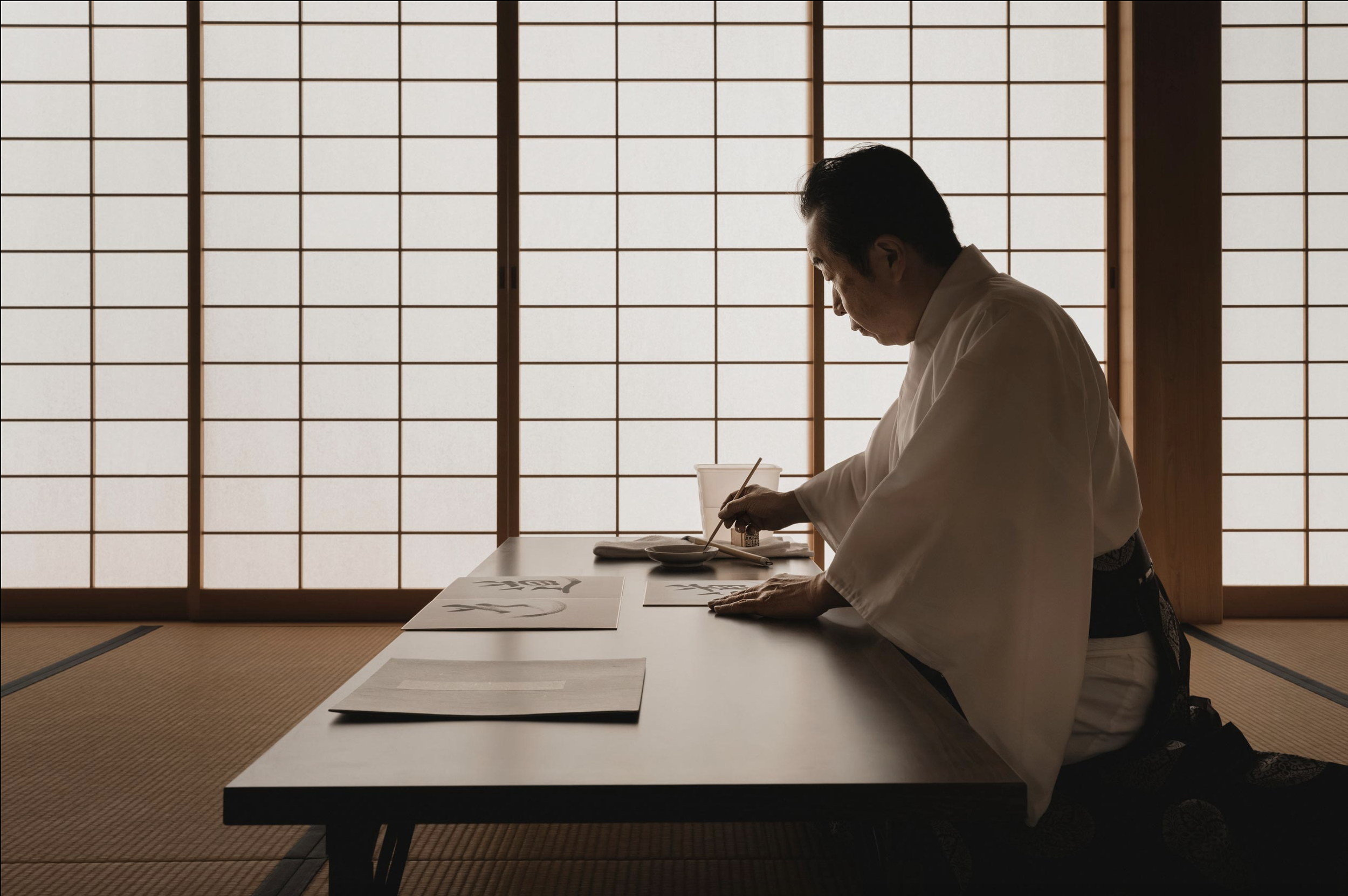 Un hombre en ropa tradicional japonesa practicando caligrafía en un ambiente de estilo japonés con puertas shoji.