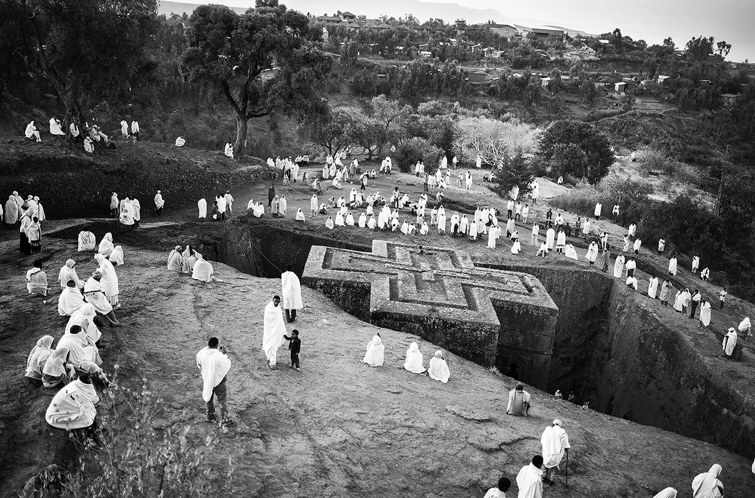 Personas vestidas con túnicas blancas en un sitio arqueológico con un posible diseño de símbolo en el suelo, con árboles y colinas en el fondo.