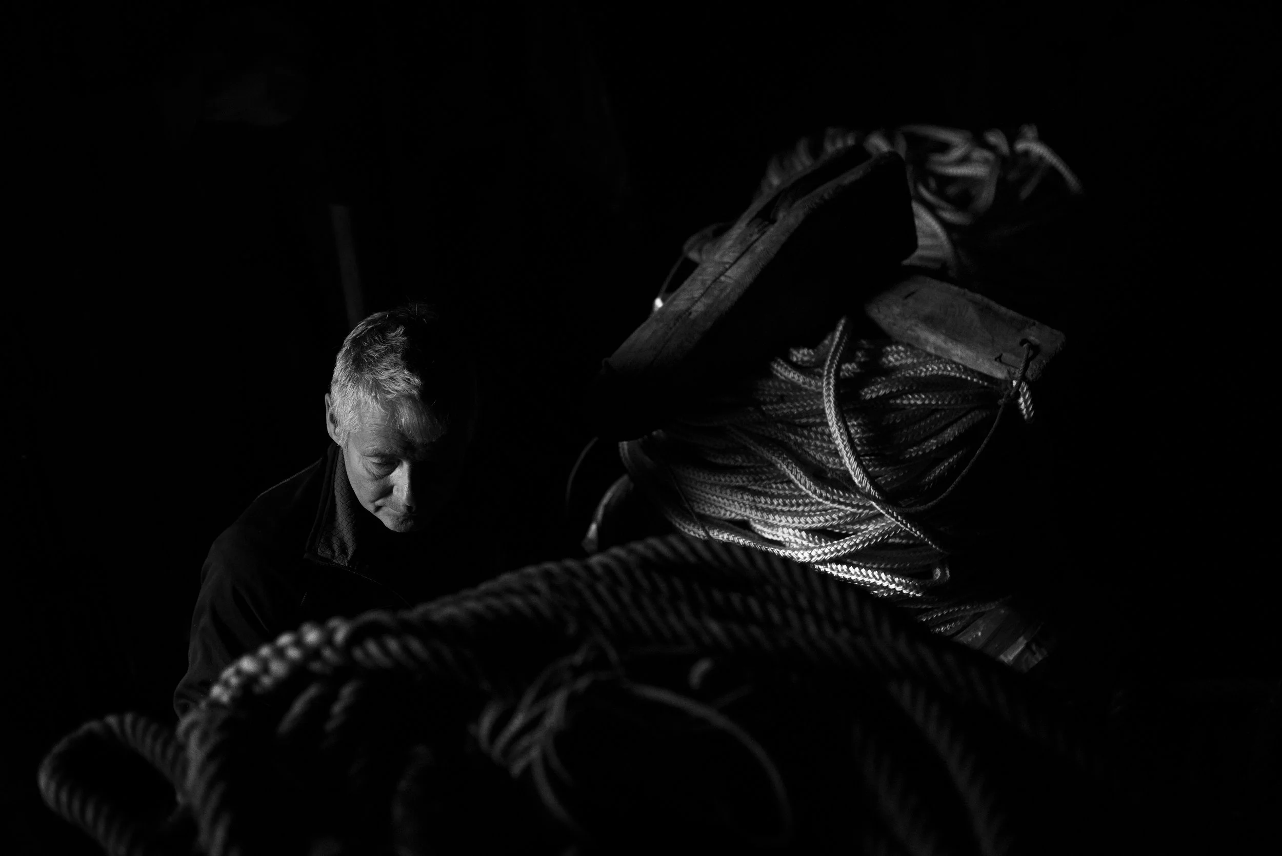 Un hombre de cabello canoso en un entorno oscuro, rodeado de cuerdas y equipos de escalada, con una expresión concentrada.