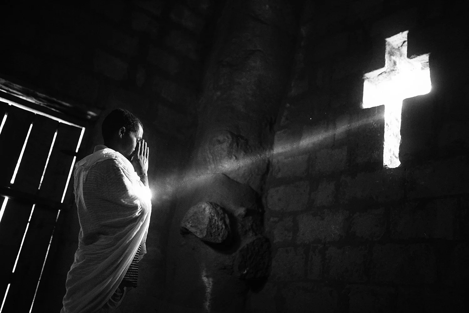 Una persona en un interior de piedra, de espaldas, en oración, con luz solar entrando por una ventana en forma de cruz.