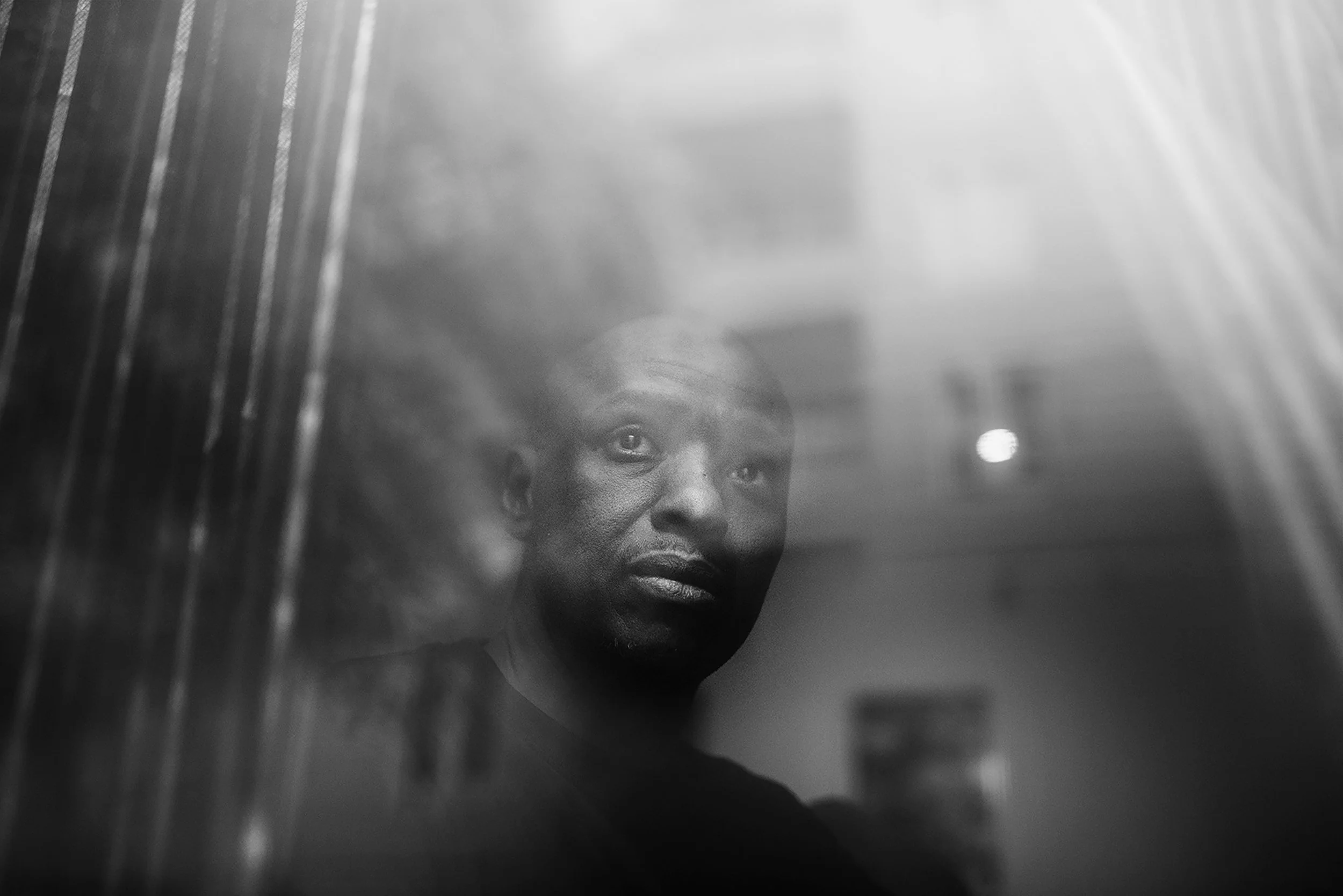 Retrato en blanco y negro de un hombre mirando a través de una ventana con reflejo, en un ambiente interior.