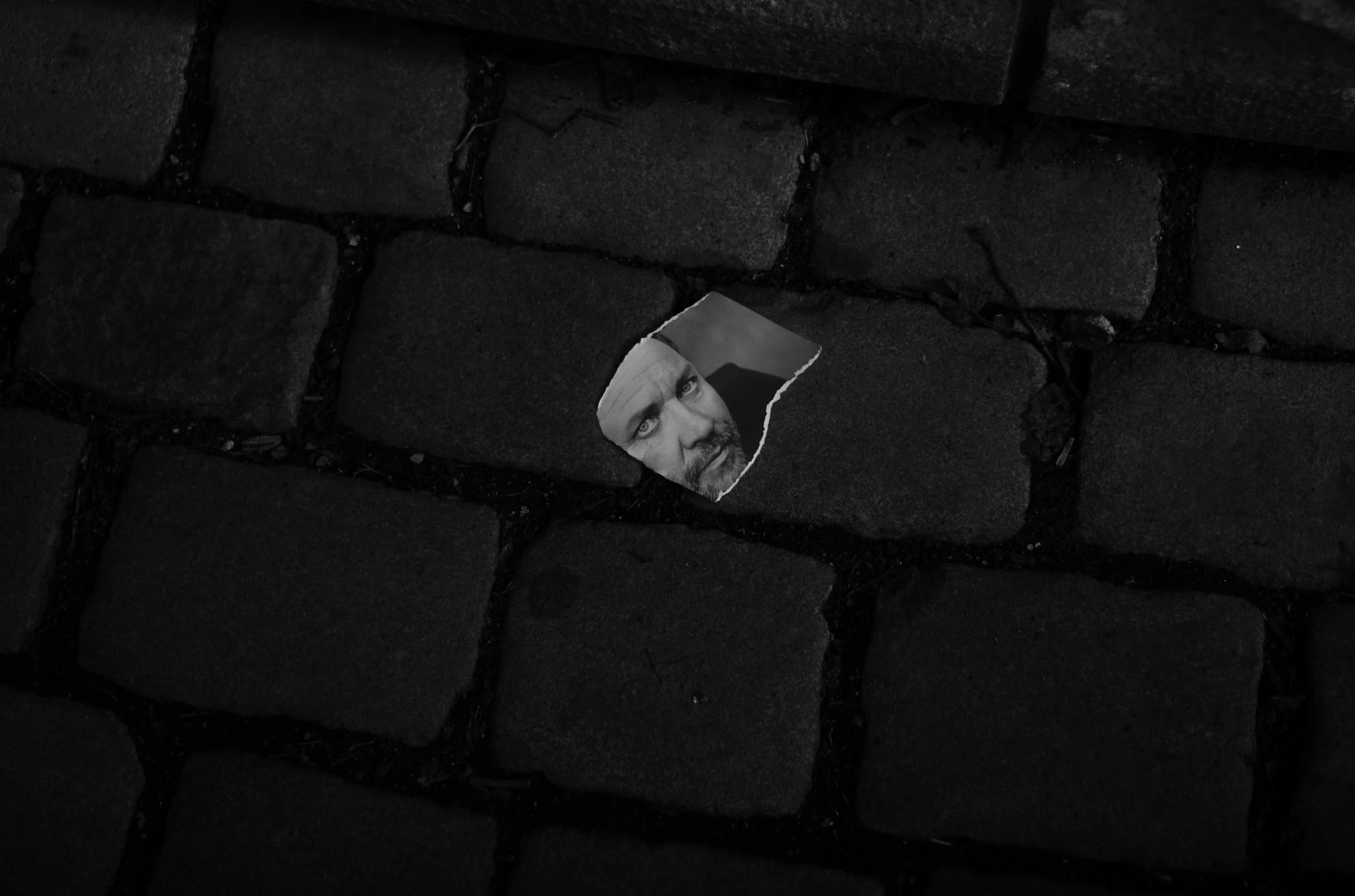 Una imagen en blanco y negro de un suelo de adoquines con un trozo de papel que muestra la cara de un hombre con barba y ojos claros.