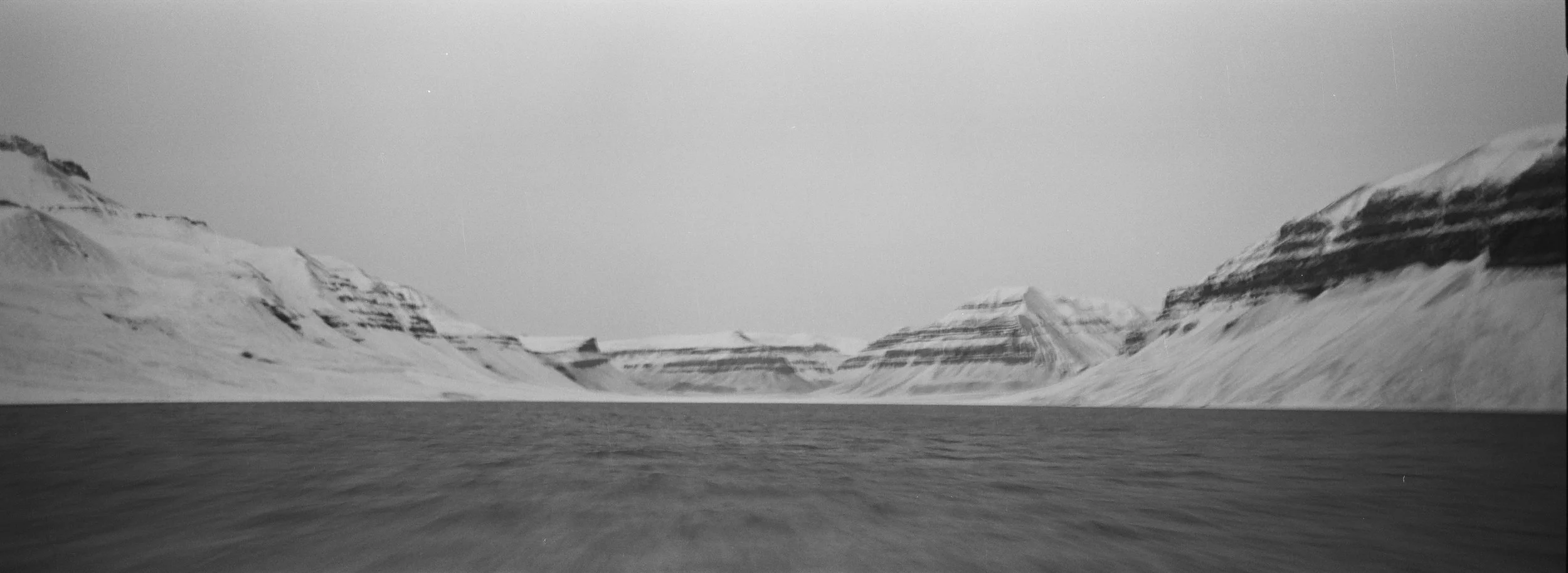 Paisaje en blanco y negro de montañas con cumbres cubiertas de nieve y un cuerpo de agua en primer plano.