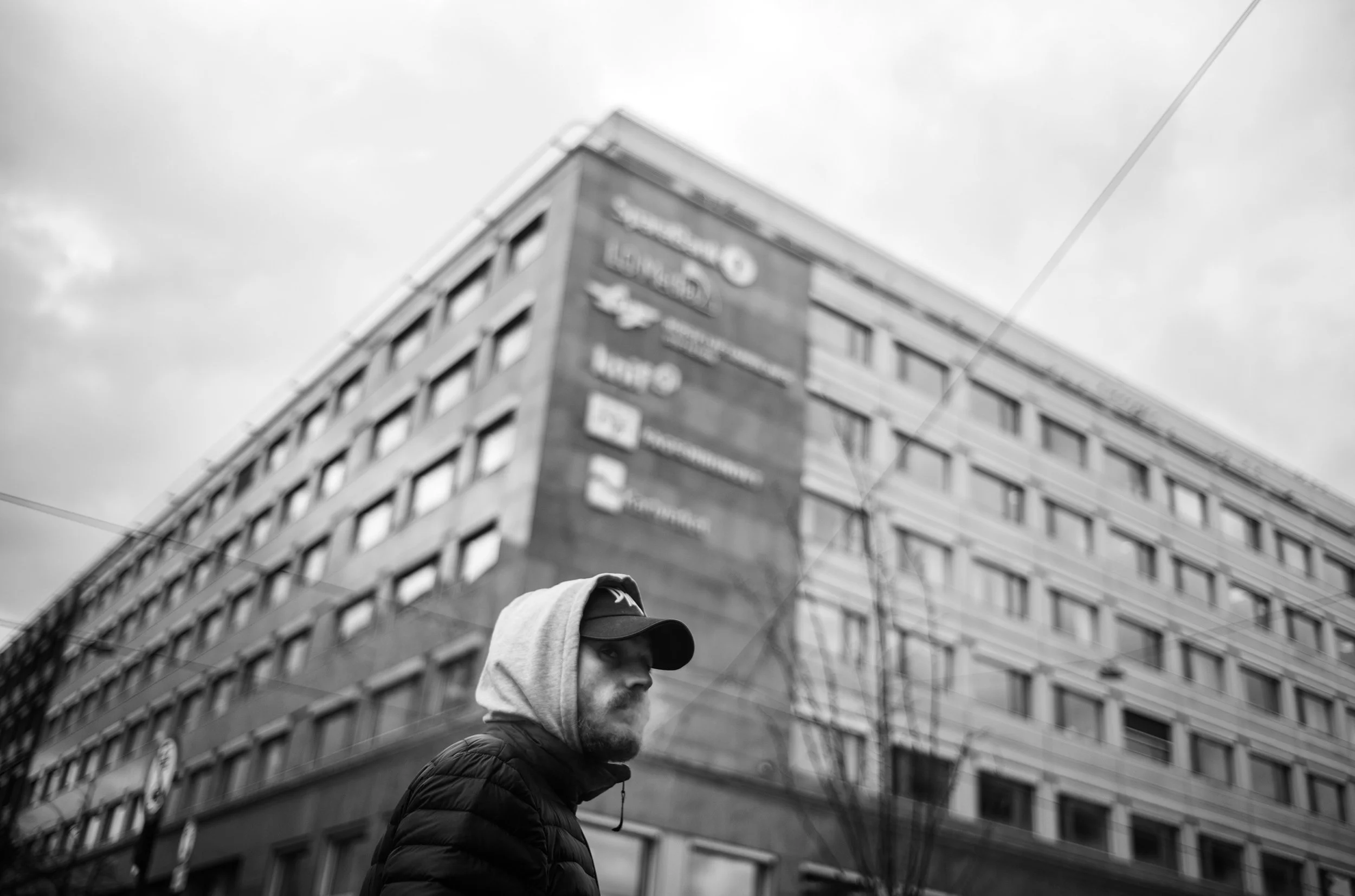 Persona con gorra y sudadera con capucha en primer plano, edificio en el fondo con anuncios en inglés, cielo nublado.