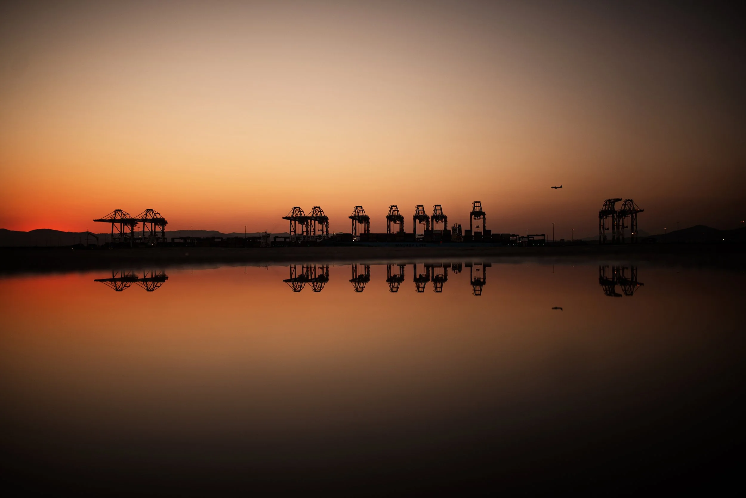 Puertos con grúas para carga y un avión en el cielo durante el atardecer, reflejados en el agua.