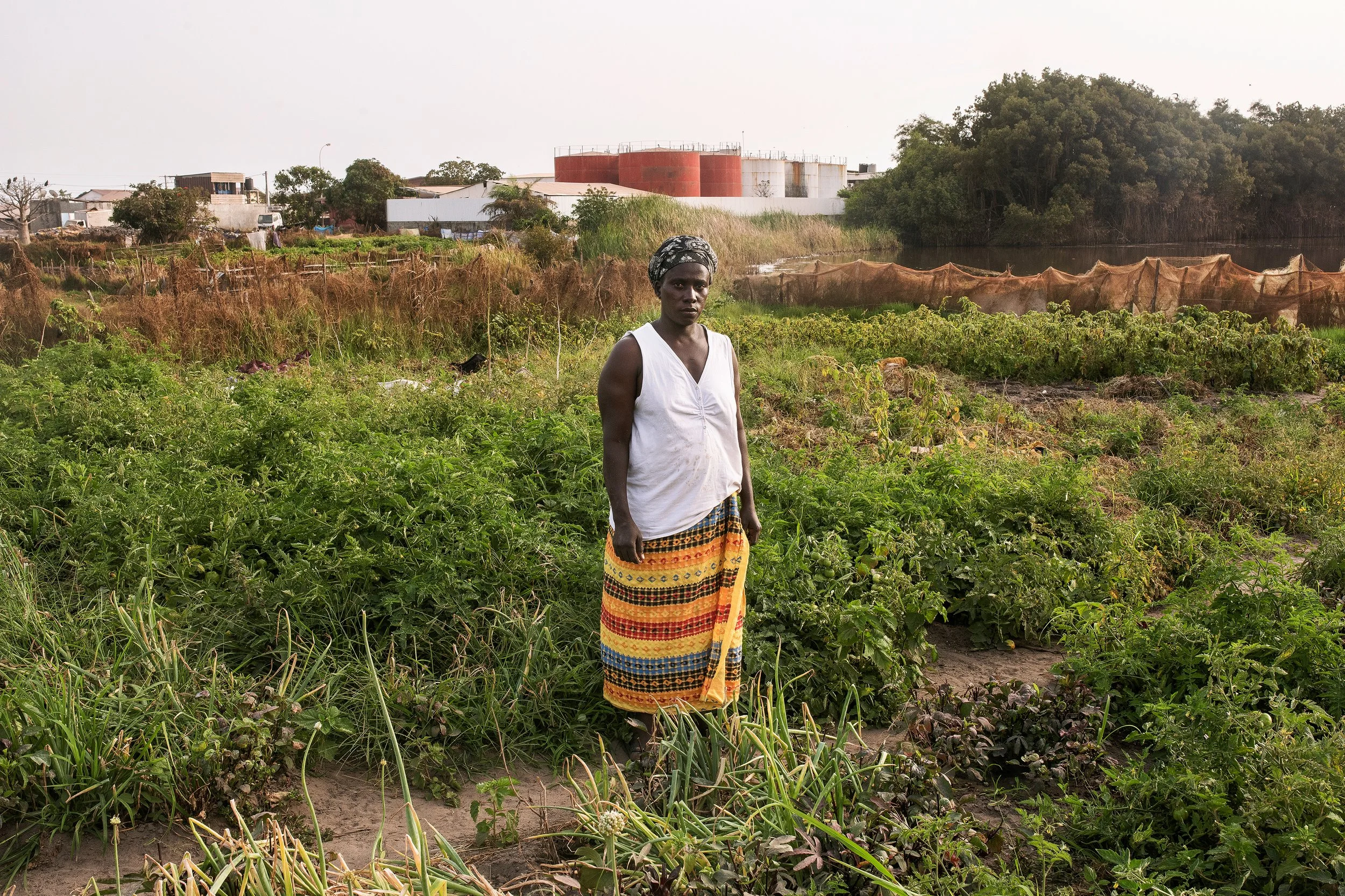 Mujer negra en un campo de cultivo con vegetación y una estructura industrial en el fondo